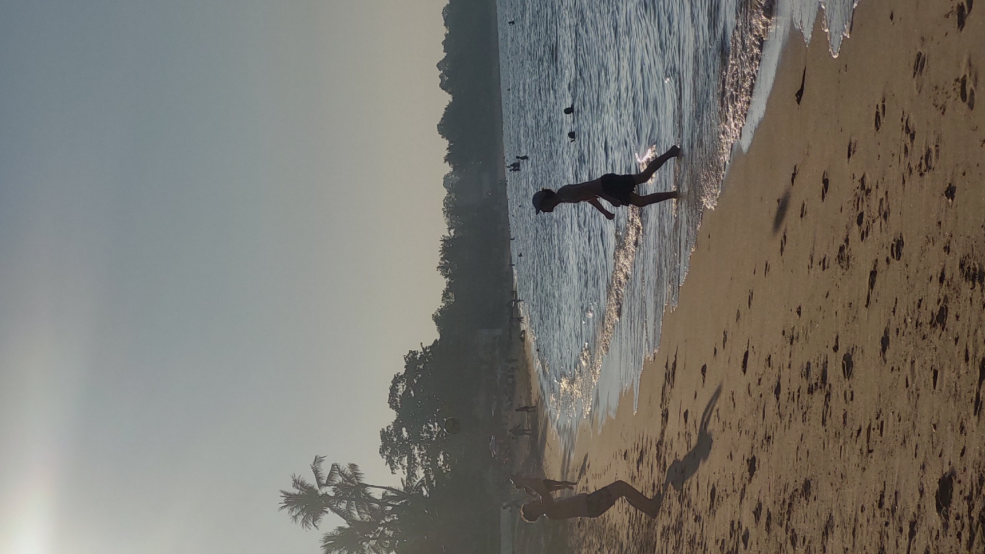 The image captures a beach scene in Baan Mae Nam, Thailand. The beach is sandy, with numerous footprints visible. The water edge is marked by small waves. A boy is shown running in the water. Other individuals can be seen in the background, both in the water and on the shore. Palm trees are located on the left side of the image. The sky is clear, suggesting a sunny day.