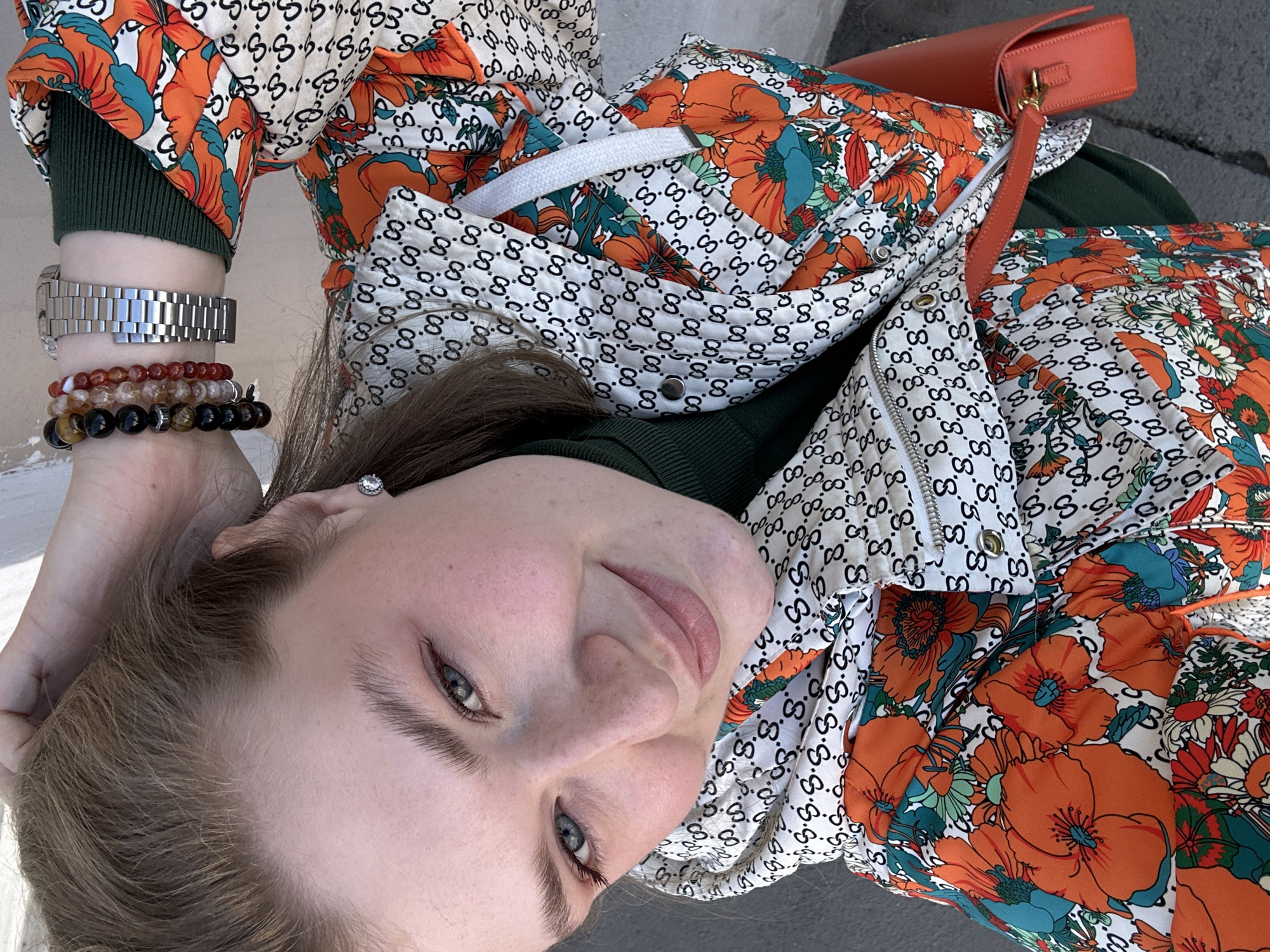 A medium-shot, low-angle view of a fair-skinned person, likely female, lying on a gray, potentially concrete, surface outdoors, possibly in the city of Kazan, Russia, inferred from contextual information. The person is wearing a green turtleneck under a jacket with a floral and logo print in shades of orange, white, and black. They have light brown hair, and a single earring is visible on their left ear. A metal watch and beaded bracelets adorn their left wrist. A red purse is visible next to the jacket. The person is looking towards the camera, and the overall composition suggests a casual, perhaps self-taken, photograph.