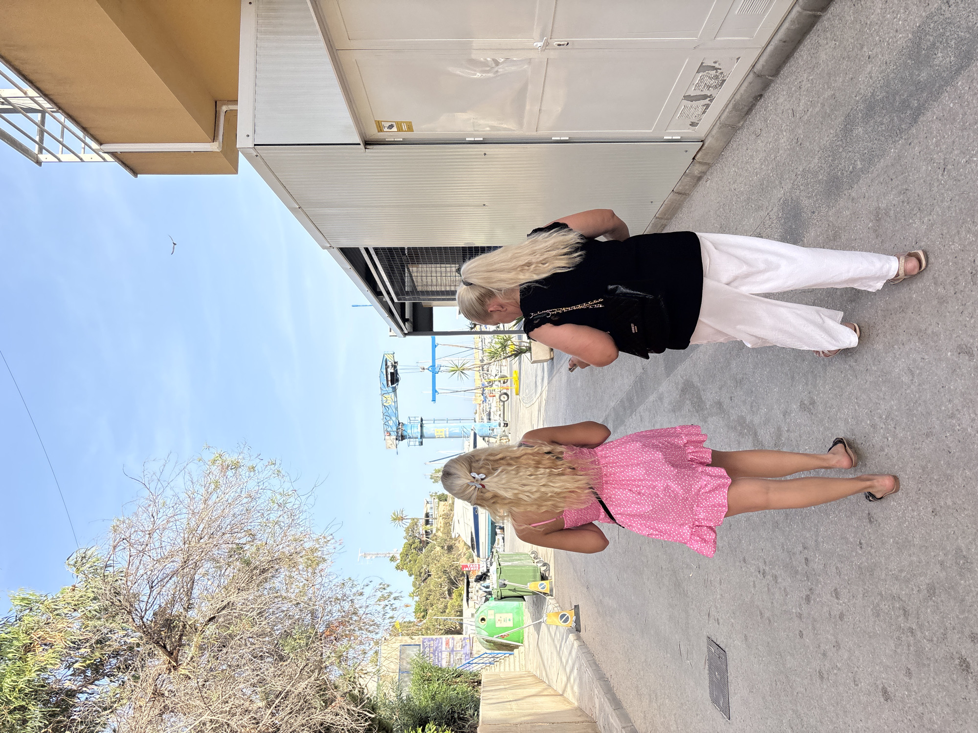 In Orihuela, Spain, two blonde women with their backs to the camera walk down a paved street lined with buildings and industrial containers. The woman on the left wears a pink polka dot dress and sandals. The woman on the right wears a black top and white pants with sandals. A white industrial building is on the right side of the image. A tree with sparse green and brown leaves is on the left, and in the background, a blue crane is visible near a body of water with boats. The sky is clear and blue.