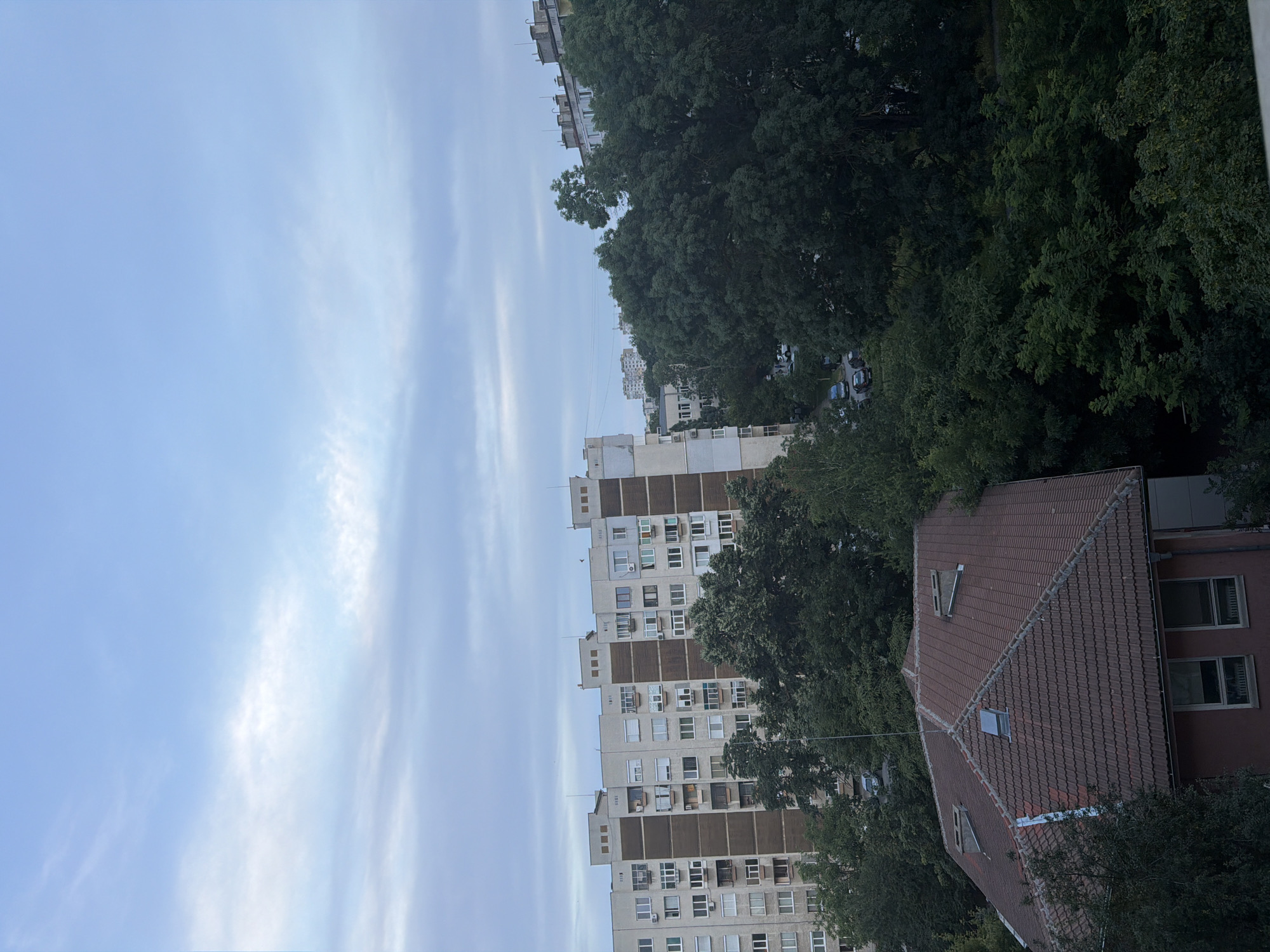 The image depicts a residential area in Ruse, Bulgaria, under a cloudy sky. In the foreground, a brown tiled roof of a building with visible dormer windows and chimneys is prominent. Behind it, a large, multi-story apartment building with a light-colored facade and darker brown vertical sections dominates the mid-ground. Numerous windows are visible on the apartment building, some with air conditioning units. Dense green trees partially obscure the lower floors of this building and extend throughout the scene, creating a lush, natural element within the urban environment. In the background, more apartment buildings and trees are visible, along with several parked cars on a street. The sky is overcast with soft, diffused light. The overall impression is of a typical, somewhat dense, residential neighborhood in Ruse.