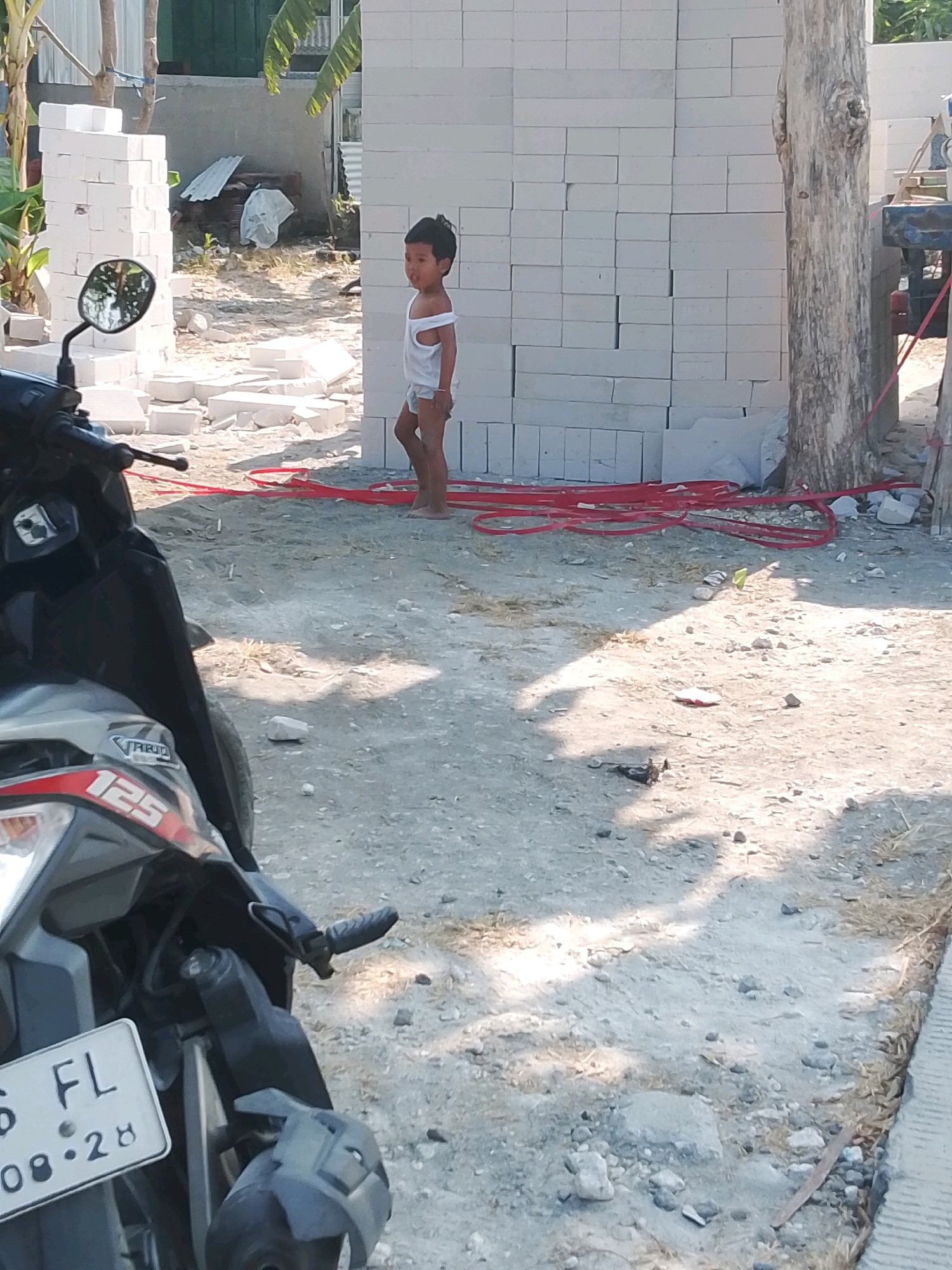 A young boy stands on a dusty construction site in Wadang, Indonesia. He is wearing a white tank top and grey shorts. The boy is looking to his right, with his mouth slightly open. The background is filled with stacks of white cinder blocks, some of which are scattered on the ground. Red piping lies on the ground in front of the blocks. To the left of the boy, a motorcycle is parked. The ground is covered in sand and small rocks, with patches of dry grass. A large tree trunk stands to the right of the boy. The scene appears to be an outdoor construction area with building materials and a motorcycle.