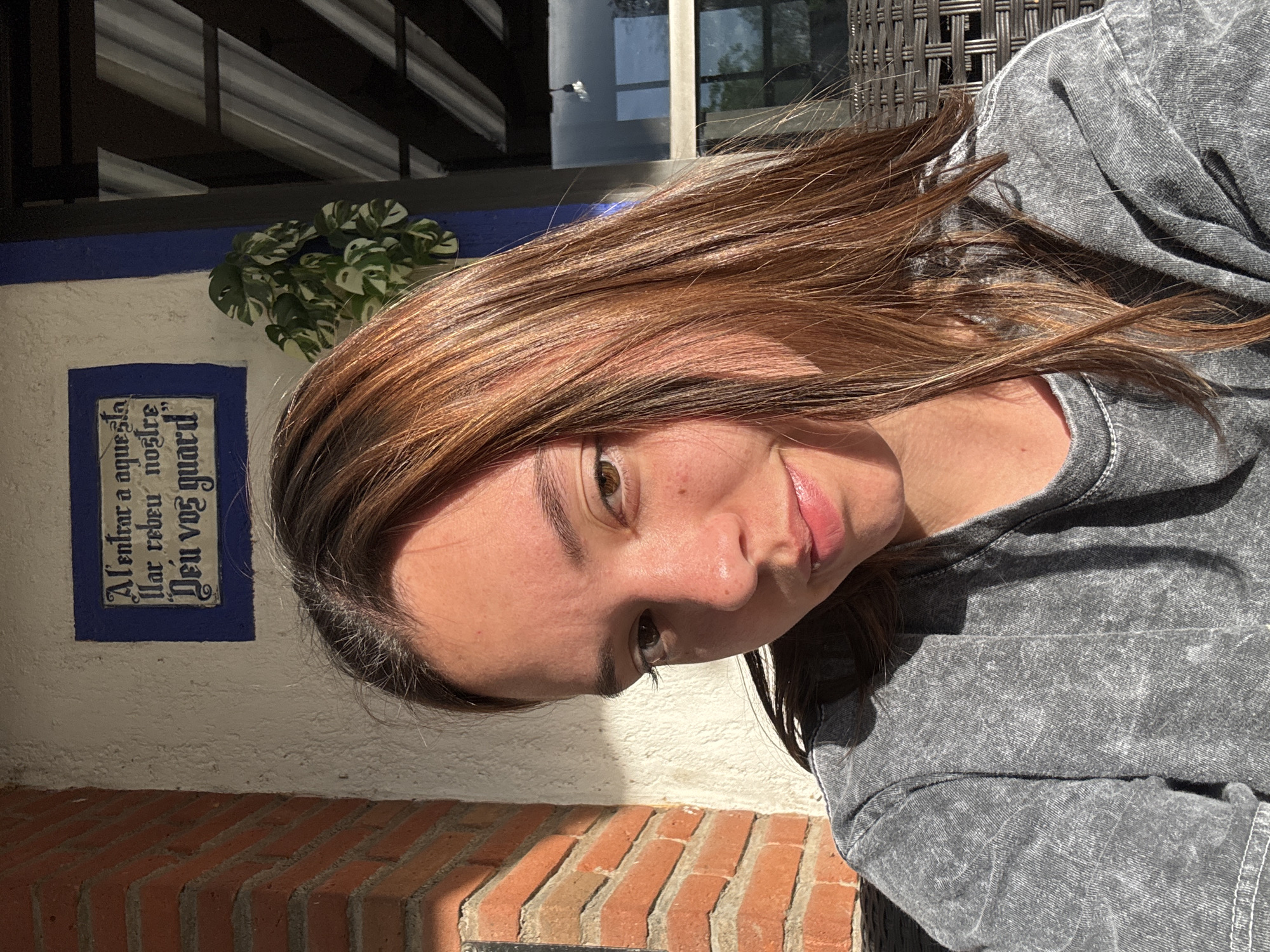 A photograph shows a woman with brown hair and a gray shirt. She is outside on a sunny day in Puebla, Mexico. Behind her is a wall, a building facade, and vegetation. The wall has a sign, and an outdoor seating area is visible at the top of the frame.