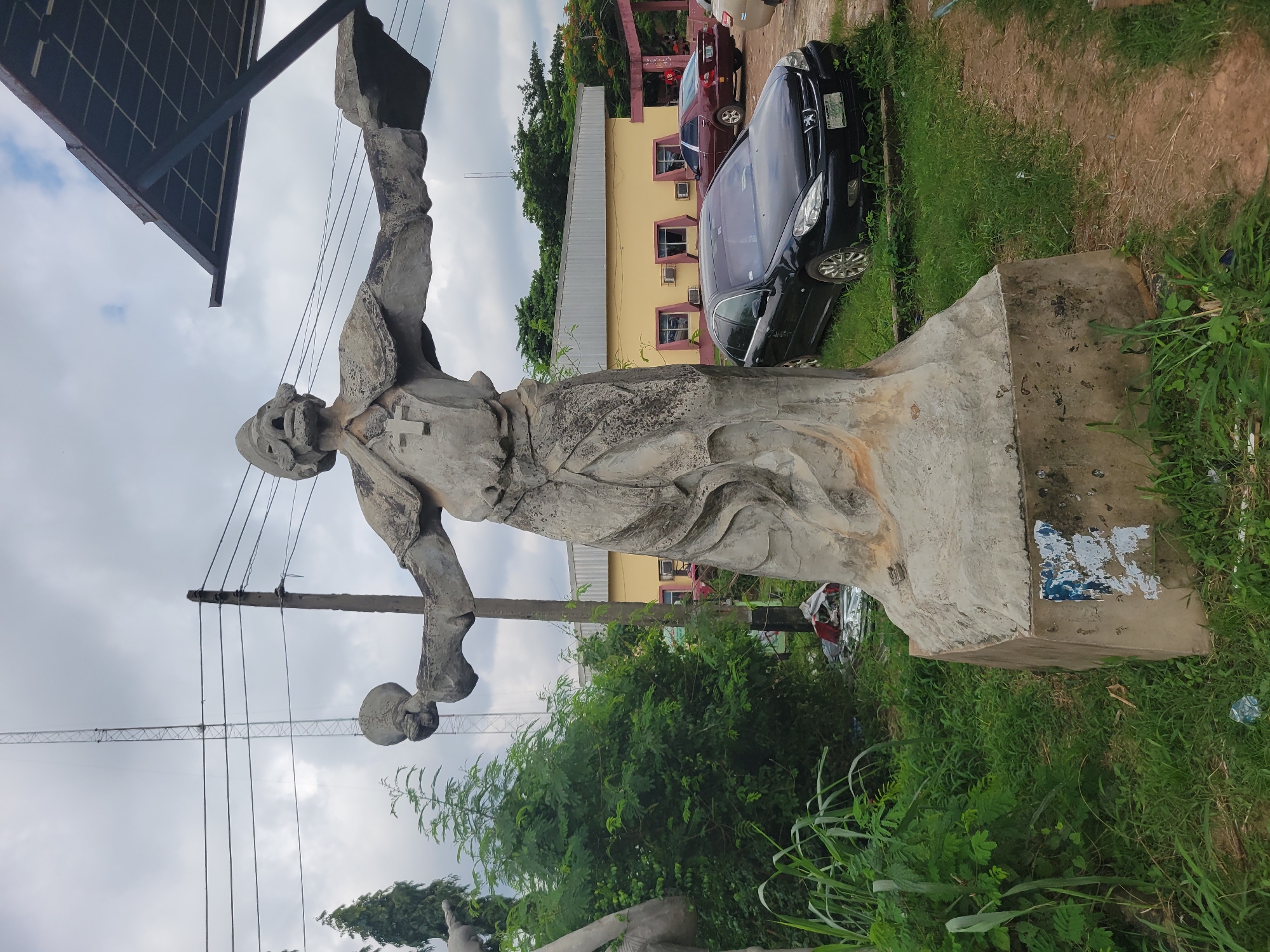 An outdoor, eye-level, full shot captures a weathered, concrete statue of a robed figure with outstretched arms, holding objects in each hand. The figure wears a tall headdress and a cross emblem on its chest. To the left of the statue, a tall transmission tower pierces a cloudy sky, with power lines strung between poles. Adjacent to the statue, a lush green bush and tall grasses grow. In the background, a yellow building with a metallic roof is visible, featuring several windows with dark frames and air conditioning units. A black Peugeot sedan is parked on a dirt path in front of the building, and a maroon sedan is parked further to the right. The scene appears to be in a semi-urban or rural setting in Egbagu, Nigeria, as indicated by the statue's style and the overall environment. The statue stands on a concrete base adorned with peeling blue and white posters. A solar panel structure is positioned above and behind the statue on the right side.