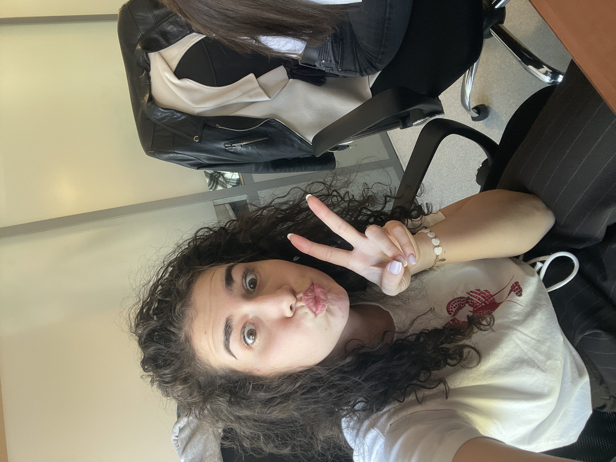 The image, rotated 90 degrees clockwise, captures a young woman taking a selfie in an indoor setting, likely an office, in Sofia, Bulgaria.
The main subject is a woman with dark, curly hair, light skin, and wide brown eyes. Her head is positioned on the left side of the frame. She is looking directly at the camera, pursing her lips in a playful kissy face, and raising her left hand to make a peace sign. She wears a white t-shirt featuring a prominent red lobster graphic on the chest. A bracelet with small, light-colored, heart-shaped beads is visible on her left wrist, and her fingernails are painted with a light, possibly pale pink or nude, polish.
In the background, on the right side of the image, a black leather jacket with metallic zippers and a light-colored (cream or white) ribbed top are draped over a black office chair. Below this, the base and chrome wheels of another office chair are visible on a light grey floor. A dark, pinstriped fabric can be seen on the far right. The overall background is a plain, light-colored wall or partition.
The activity is a playful selfie, evident from her pose and expression. The lighting suggests daytime. No discernible text is visible on any objects.