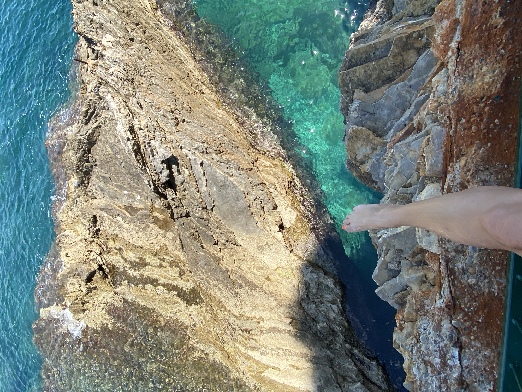 The image shows a view from above a rocky cliff into the turquoise waters below, likely near Budva, Montenegro, based on the characteristic coastal rock formations and water color of the Adriatic Sea in that region. A person's foot extends over the edge of the cliff. The rock formations display a layered, sedimentary structure with colors ranging from brown to gray to tan. The water is clear, revealing rocks and the sea bottom. The lighting suggests a sunny day, with sunlight reflecting off the water's surface. The angle indicates the photo was taken directly downward.