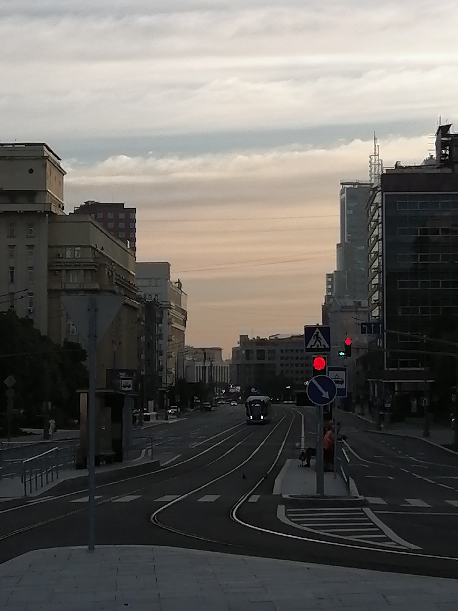 The image shows a street scene in Moscow, Russia, during what appears to be dusk or early morning, given the soft lighting and the sky's colors. The street is wide with tram tracks running down the center, curving to the right in the foreground. On the left side of the street, several multi-story buildings are visible, some with neoclassical architectural elements. On the right side, modern high-rise buildings with glass facades dominate the skyline.
Traffic signals are present, with a red light illuminated on a signal pole in the middle right of the frame, above a pedestrian crossing sign. A blue directional sign with a white arrow pointing left is also visible. A tram is moving away from the viewer down the tram tracks in the distance.
Several people are on the sidewalk near the tram tracks. On the right, a person wearing an orange vest is standing near a pole, and another person is seated on a bench. A pedestrian crossing is marked on the road. The overall impression is a busy urban environment with a mix of old and new architecture.