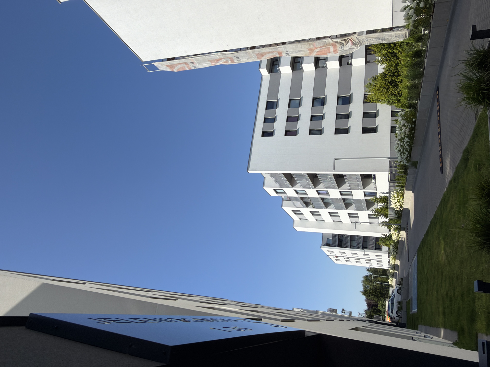 The image depicts a modern residential complex in Poznan, Poland, under a clear blue sky. On the left, a building with a dark facade and a light-colored, angled side is visible. Further into the scene, a row of white apartment buildings with grey accents and balconies stretches across the frame. These buildings have multiple floors and numerous windows. A large white banner with red lettering is attached to scaffolding on the rightmost building, obscuring part of it. In the foreground, a well-maintained lawn borders a paved area where cars are parked. A speed bump is visible on the paved road. Lush green trees and white flowering bushes line the walkway, creating a neat and organized urban landscape. The scene is bathed in bright sunlight, indicating a clear day.