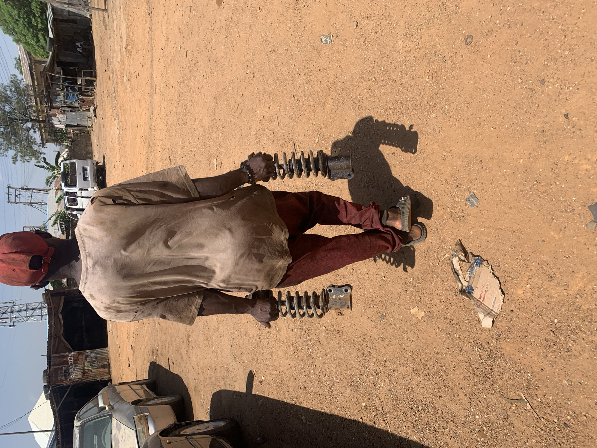An individual wearing a red cap, tan shirt, and red pants stands on a dirt/sand surface, holding two car shock absorber struts with springs in their hands. The environment is an outdoor location, likely a street or roadside in Rafin Par, Nigeria, indicated by the presence of local buildings, vehicles (cars and a white van), and cardboard with the text "Nigeria PLC". The person is facing away from the camera. A partially crumpled cardboard box is on the ground nearby, along with the person's shadow.