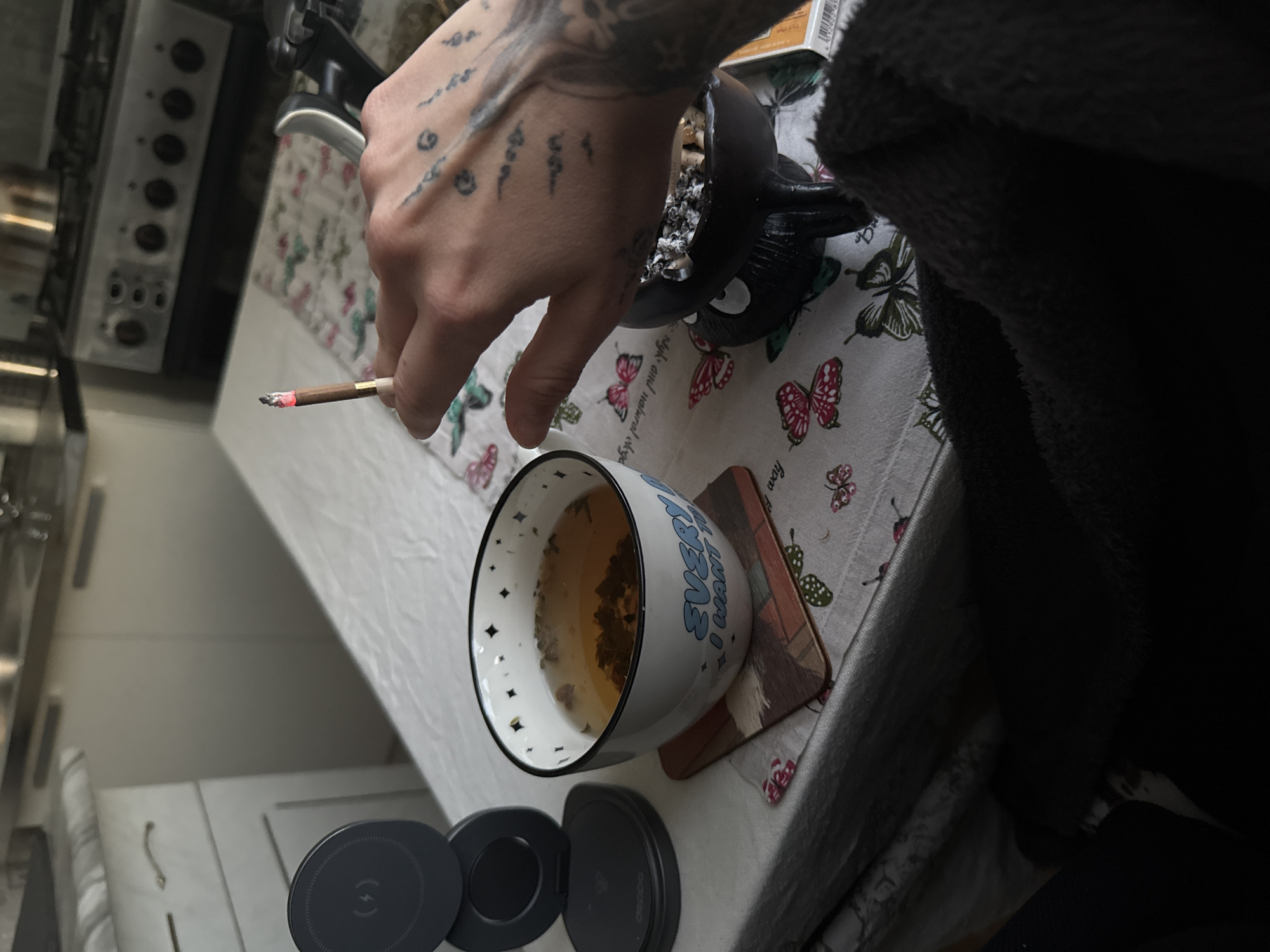 The image, taken in a domestic setting, likely a kitchen or dining area, depicts a close-up of a person's hand holding a lit cigarette or joint, positioned above a table with various objects. The scene suggests a moment of relaxation.
The subject is primarily a tattooed left hand and forearm. The hand features extensive black ink tattoos, appearing to be in a script or symbolic style, possibly Sak Yant or similar, with multiple distinct patterns visible across the knuckles and back of the hand. The person is holding a lit, filter-tipped cigarette or joint, with a glowing red cherry at the tip, indicating it is actively burning.
On the table, covered by a white tablecloth adorned with numerous colorful butterfly patterns (pink, red, green), several items are present. A black, character-shaped ashtray, resembling a stylized creature like Totoro, is filled with cigarette ashes and butts. Next to it, a white ceramic mug, decorated with black speckles around the rim, contains a dark-colored liquid, likely tea, with visible tea leaves or herbs. The mug has blue text on its side, partially visible, which reads "EVERYTHING I WANT". Underneath the mug is a small, brown coaster with a textured surface.
In the lower left foreground, two dark grey circular objects are visible, one of which displays a wireless charging symbol (a circle with a stylized 'S' or lightning bolt inside), suggesting it's a wireless charger. The background reveals parts of a kitchen, including a silver stove or oven with several control knobs on the side, and white cabinets. A dark, possibly furry, fabric object (clothing or blanket) partially obscures the right side of the image.
Visible text on the tablecloth includes fragmented phrases like "light and natural" and "how". The tattoos on the hand are not clearly legible but appear to be script or symbols. A barcode and some numbers are faintly visible on a white rectangular object in the upper right, partially obscured by the dark fabric. The overall impression is a casual, indoor scene. No specific time of day is indicated by the lighting. While the context provides Saint Petersburg, Russia, there are no specific visual cues within the image to confirm this location.