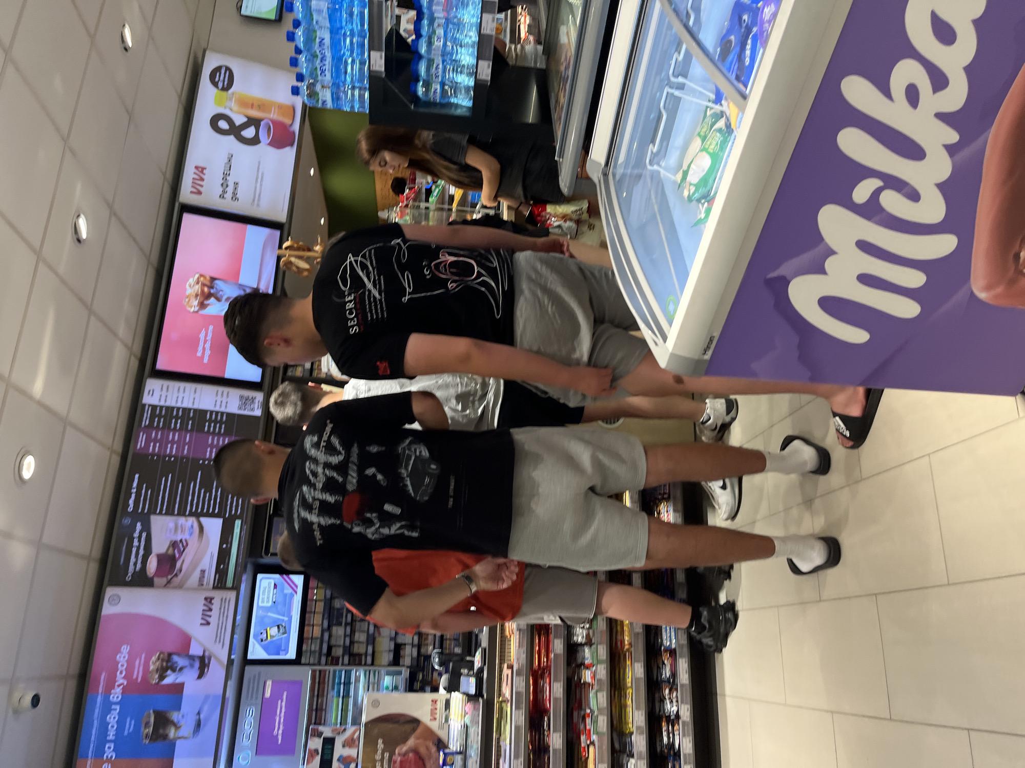 The image depicts the interior of a convenience store, possibly in Popovitsa, Bulgaria, given the Cyrillic text on the advertising displays. A "Milka" branded refrigerated display case is prominent in the foreground. Several customers are present, including at least two men wearing black t-shirts and grey shorts who are standing near the display case. One woman with long brown hair is visible in the background, near bottled water. Aisles of shelving stocked with various products stretch into the distance. There are video displays above advertising drinks and coffees.