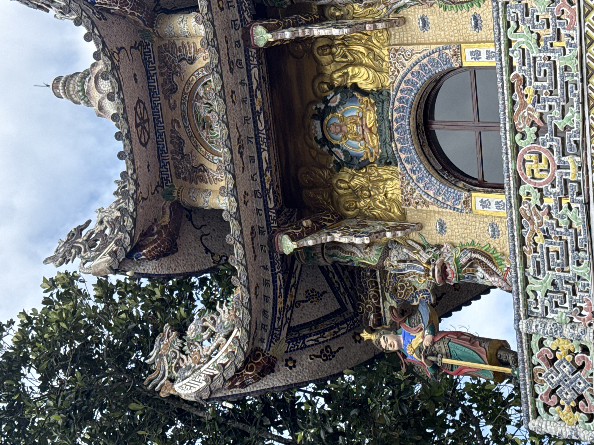 This image captures a section of an ornately decorated Buddhist temple or pagoda, located in Xuân Thành, Vietnam, under an overcast sky with dense green foliage visible on the left.
The structure features multiple tiers, primarily composed of intricate mosaic tile work in earthy tones, blues, greens, and whites. At the apex, a multi-tiered spire with diminishing rings is visible against the sky. The curved roofline below the spire is adorned with stylized dragon figures and geometric patterns, and its underside displays a border of Buddhist script.
Below this, the main facade presents a prominent golden relief centered with a seated Buddha figure on a lotus throne, enclosed in a round halo, flanked by multiple standing Buddha or Bodhisattva figures, also with halos. Below this relief is an arched window with dark panes, framed by mosaic patterns and vertical panels bearing Chinese characters.
On the lower visible level, a standing guardian figure is positioned on the left, clad in colorful garments (blue, green, red) and a golden helmet, holding a staff or sword. To its right, an elaborate dragon head sculpture emerges from the building's support. The railing or balustrade beneath these figures is richly detailed with geometric patterns, including a left-facing swastika symbol (Buddhist symbol of auspiciousness), all executed in mosaic tiles. The building's surface is consistently covered in small, multicolored mosaic tesserae.
