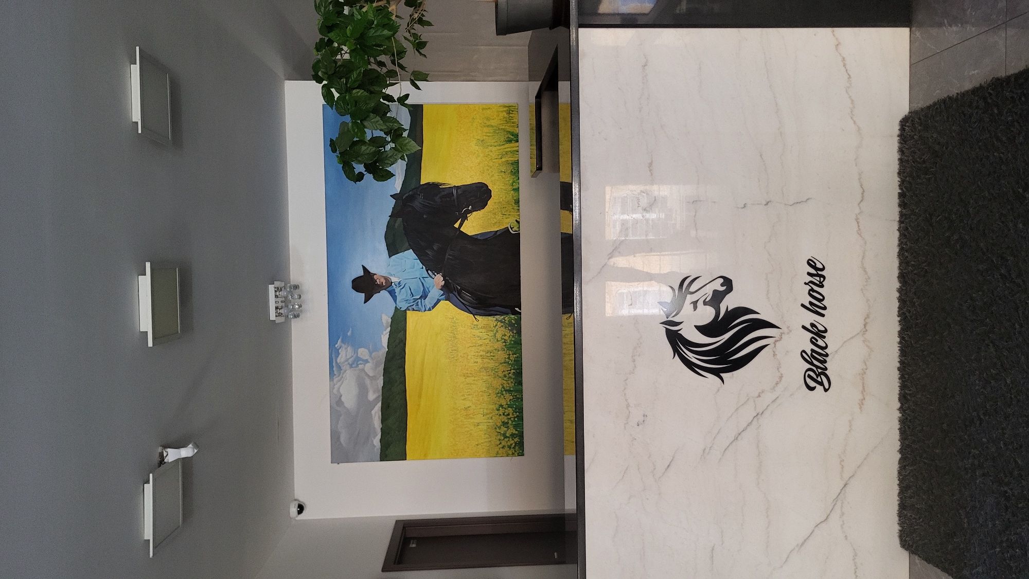 A painting of a cowboy on a black horse in a yellow field hangs above a reception desk. The reception desk has a marble-like finish and a logo of a black horse with the text "Black horse" below it. A dark grey rug is in front of the desk. The ceiling has three light fixtures, one of which appears damaged with a white object hanging from it. A security camera is visible on the left side of the wall. A potted plant is on the right side of the desk. The overall scene suggests the reception area of a business or establishment, possibly related to horses or equestrian activities.