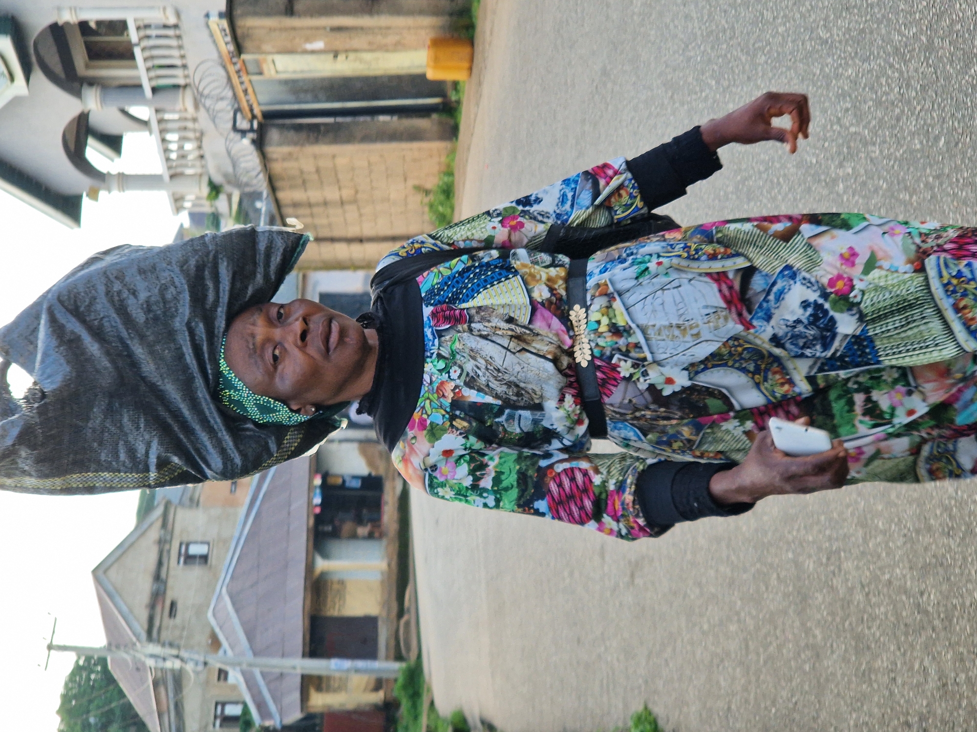 A Nigerian woman, likely from Kabong, Nigeria, carries a large black bag balanced on her head. She is walking down a paved street lined with buildings and vegetation. The woman is wearing a colorful, patterned dress and a green headscarf. She is holding a white smartphone in her right hand. The background shows residential buildings with varying architectural styles. The scene appears to be captured during the day.