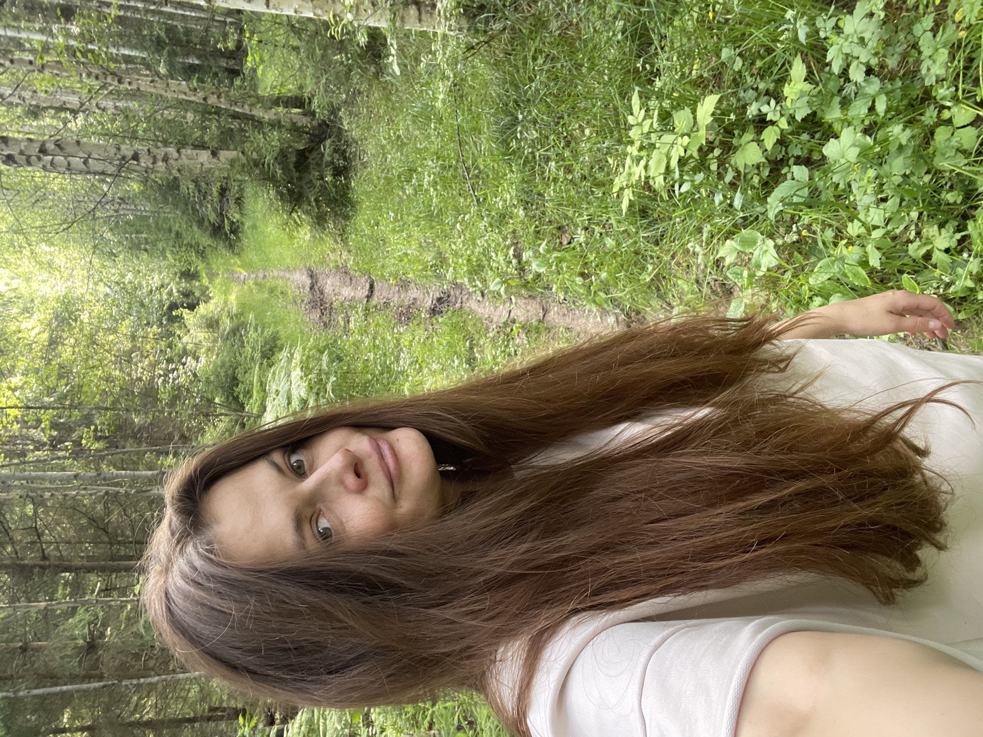 This is a portrait of a young woman with long, dark brown hair, taken outdoors in a forest setting. She is positioned in the foreground, with her head tilted and her gaze directed towards the camera. Her expression appears relaxed and perhaps a little curious. She is wearing a light-colored top.
The background reveals a wooded area with a path winding through it. The path is somewhat muddy and appears to be a trail. The trees on either side of the path are tall and slender, with some appearing to be birch trees due to their distinctive bark. Lush green foliage and grass surround the path, indicating a natural and somewhat wild environment. The lighting suggests it could be daytime, possibly a cloudy day given the diffused light, or late afternoon.
The overall impression is of a candid moment captured during a walk or exploration in a natural landscape. There is no visible text in the image. The location context provided, Коротиха, Russia, suggests a rural or natural setting within that region.