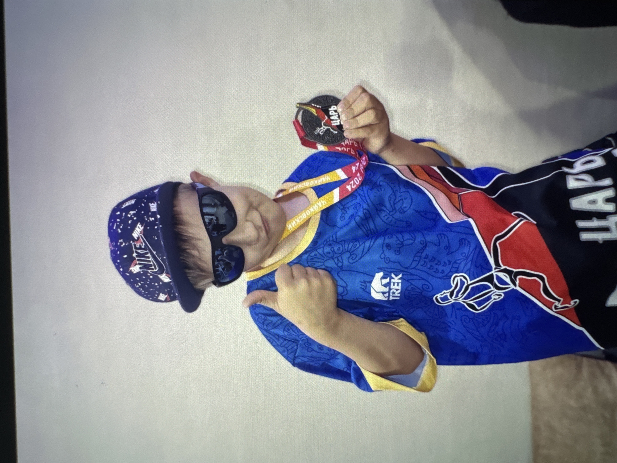 This media file captures a young individual, likely a child or pre-teen, proudly posing with a medal. The subject is seen from the chest up, wearing a vibrant blue and black sports jersey with yellow trim and intricate patterns. A white "TREK" logo is visible on the blue section of the jersey. They accessorize with a dark blue baseball cap speckled with white, featuring a prominent "NIKE" logo on the front, and dark, reflective sunglasses.
In their right hand, the individual holds up a dark-colored medal, suspended by a red and black ribbon. The medal clearly displays the Cyrillic word "ЦАРЬ" (TSAR, meaning 'King' or 'Emperor') and an emblem resembling a bird of prey. The medal's ribbon also shows "2024" and "НАЙМКОВСКИИ" (NAYMKOSKII), which could refer to a name or location. With their left hand, the subject makes a gesture resembling a thumbs-up or "shaka" sign, adding to their confident demeanor.
The activity depicted is the celebration of an achievement, presumably winning the medal. The subject appears proud and confident. The setting is indoors, with a plain, light-colored wall serving as the background. There are no specific cues for time of day or weather. The visible Russian text on the medal and ribbon is consistent with the provided location context of Chaikovsky, Russia.