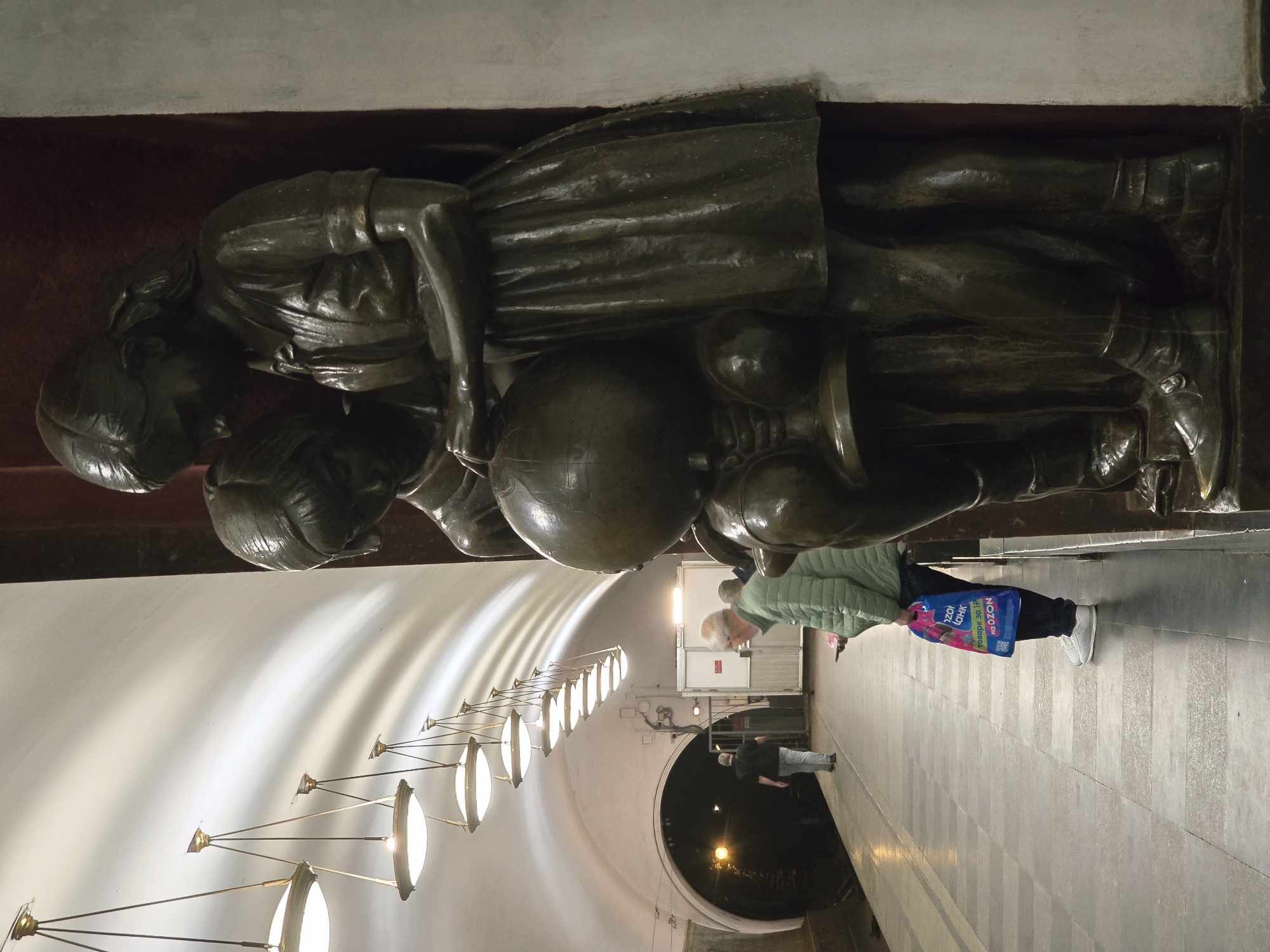 The image shows a bronze sculpture of two children playing with a globe, mounted on a wall in what appears to be a subway station. The location is Unknown, Unknown. The sculpture is above eye-level and the children are depicted in dresses. One child is leaning over the globe while the other is supporting it. Below the sculpture, the subway station has an arched ceiling with a row of evenly spaced, round, illuminated lights extending into the distance. A person in a light green jacket, dark pants, and white shoes stands on the subway platform, holding a blue and pink shopping bag. Another person in dark clothing is seen further down the platform, near the subway tunnel entrance. The platform floor is made of grey stone tiles arranged in stripes. The overall lighting is bright from the overhead lamps, contrasting with the darkness of the subway tunnel entrance.