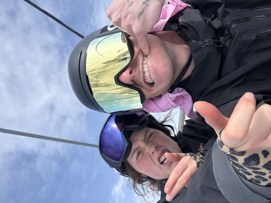 A close-up selfie depicts two individuals on a ski lift within the ski area of Le Monêtier-les-Bains, France. The subject on the left, identified as female, has reddish-brown hair and freckled skin. She wears a black ski helmet and dark goggles with a purple reflective lens. Her mouth is open with teeth visible in a grimace. Her left hand, partially covered by a leopard-print patterned garment, extends two fingers (index and middle) towards the camera. The subject on the right, identified as male, wears a black ski helmet, large gold-mirrored Oakley goggles, and a pink bandana visible around his neck/chin area. He has a light brown mustache. His left hand, which displays finger tattoos, manipulates the corner of his mouth downwards, exposing his upper teeth in a strained expression. Both individuals wear black ski jackets. A thick steel ski lift cable is visible diagonally across the upper portion of the frame, set against a partially cloudy blue sky.