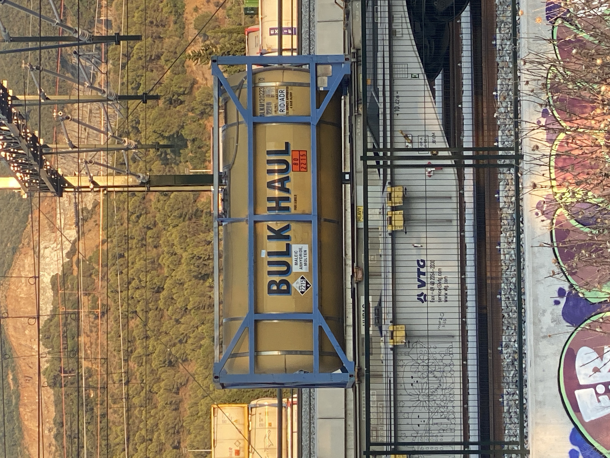 The image shows a yellow tank container labeled "BULK HAUL" on a train car in La Llagosta, Spain. The container is enclosed in a blue metal frame. There are other train cars and cargo visible in the background, along with overhead power lines and a wooded hillside. The train appears to be stationary on a railway track. The scene is lit by sunlight, casting shadows and highlighting details of the industrial setting. The graffiti on the lower concrete wall suggests an urban or semi-industrial area.