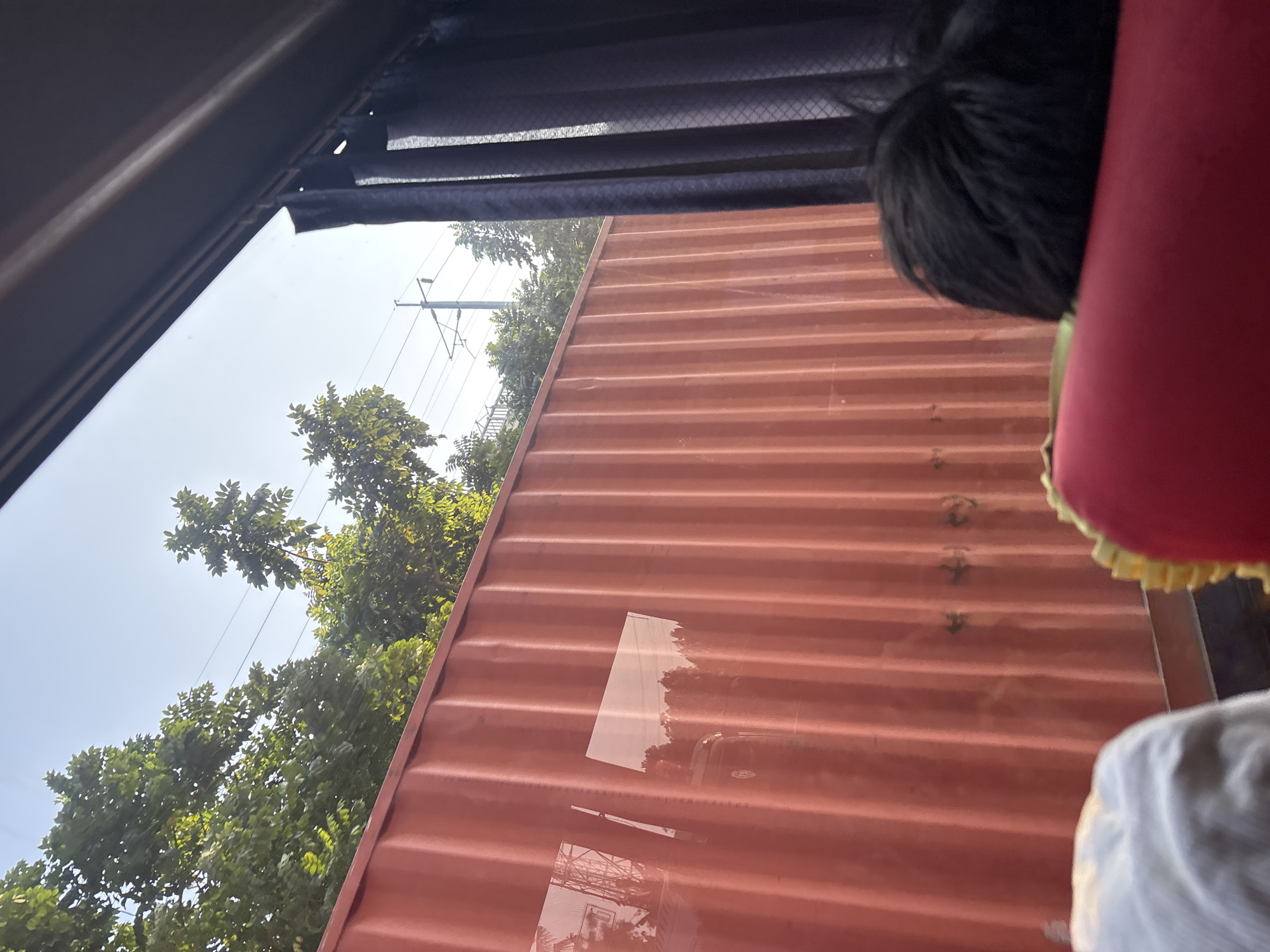 The image shows a view from the window of a vehicle, likely a bus, looking out at a bright, clear sky and lush green trees. A large, corrugated, red shipping container dominates the middle ground, reflecting some of the surrounding scenery. To the right, dark fabric curtains obscure part of the window. In the foreground, the back of a person's head with dark, messy hair is visible, as is a portion of a red seat with a yellow trim, suggesting the perspective of a passenger. The scene appears to be set in a semi-urban or roadside environment, with hints of infrastructure like power lines and poles visible between the trees. The overall impression is one of travel during daylight.