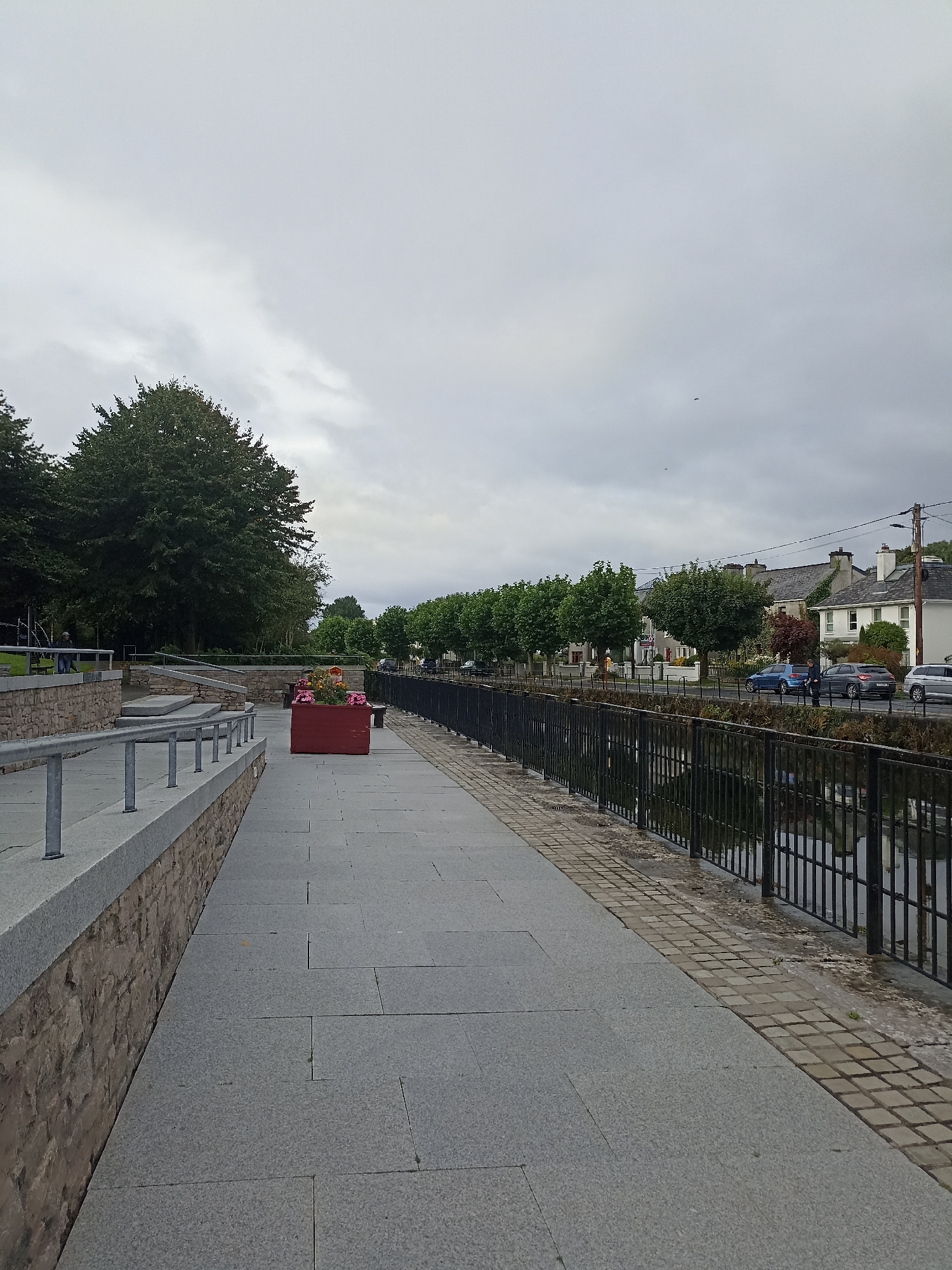The image depicts a paved walkway along a canal or river in Galway, Ireland. On the left, a tiered stone wall with metal railings overlooks the walkway. A red planter box containing flowers sits on the walkway. Beyond the walkway, a black metal fence separates the path from the water. Across the water, a row of trees lines the street, behind which are residential buildings with pitched roofs. Cars are parked and driving along the street. The sky is overcast and gray. The scene appears to be a public area designed for pedestrian access and viewing of the waterway and surrounding town.