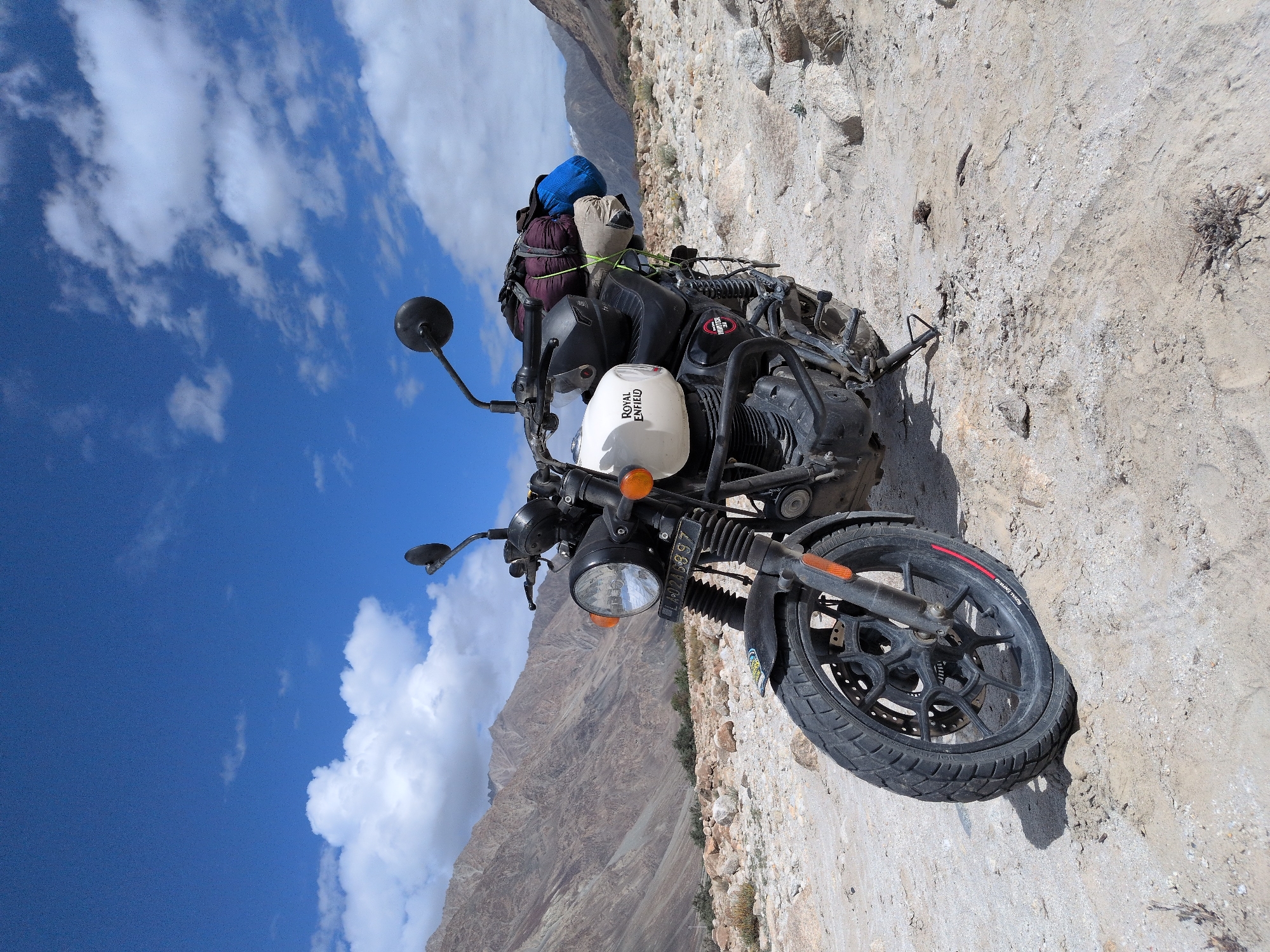 A Royal Enfield motorcycle is parked on a rocky, sandy surface with mountains in the background. The sky is blue with white clouds. The motorcycle is white with "Royal Enfield" written on the fuel tank. It has a black frame, black wheels, and a black seat. There are bags strapped to the back of the motorcycle. The license plate reads "LA-02A-5897". The scene appears to be in a remote, mountainous area, possibly in the vicinity of Sasoma, India, given the terrain and vegetation.