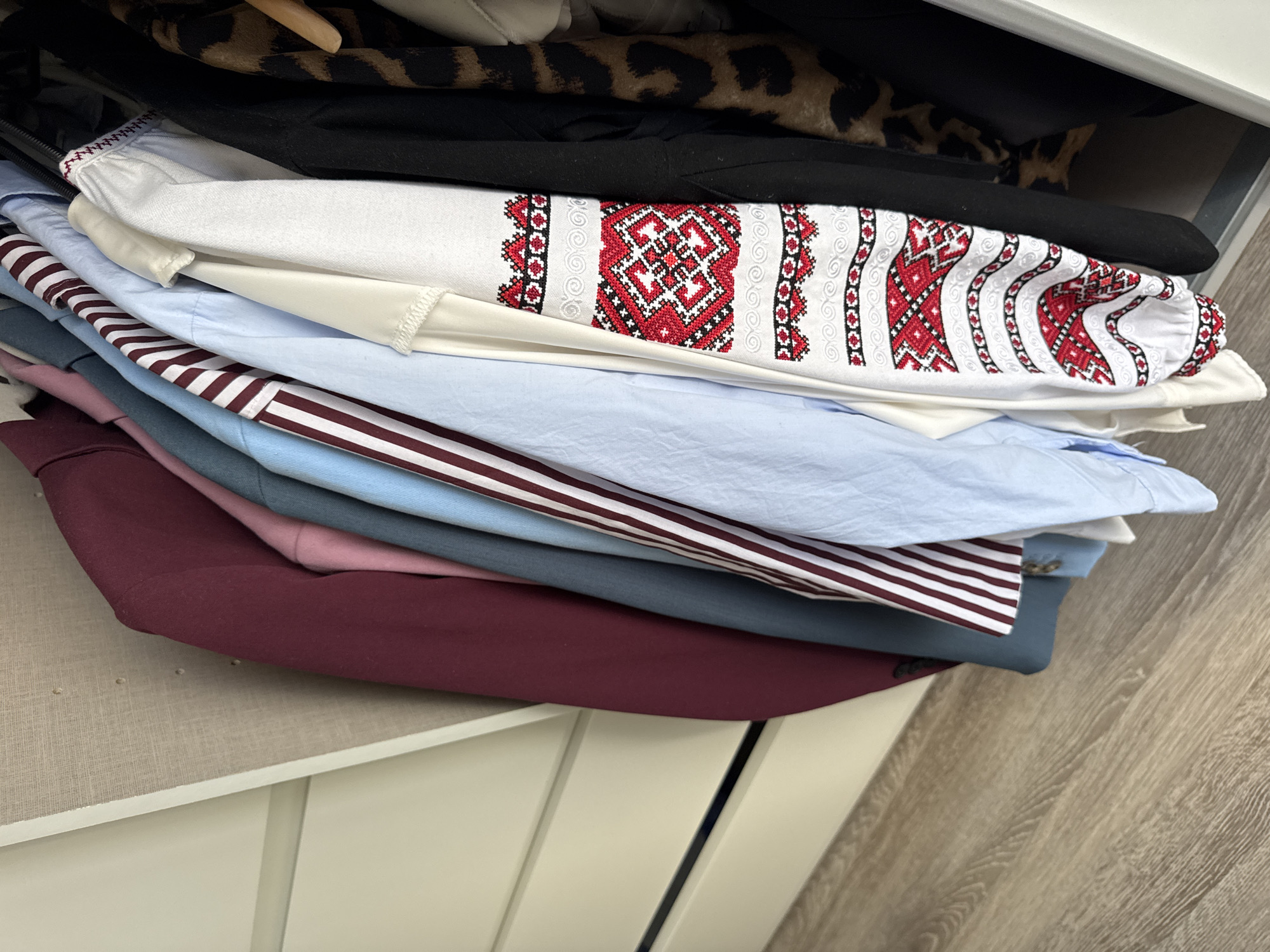 A closet in Höhenkirchen-Siegertsbrunn, Germany, contains a collection of garments hanging on a rod. The clothing includes a burgundy jacket, a pink shirt, a blue shirt, a striped shirt with burgundy and white stripes, a white shirt with red embroidered details, a black garment, and a leopard-print garment. The closet interior is visible, including a pale-colored wall and drawers. The floor is made of light-colored wood planks. The lighting suggests an indoor, daytime setting.