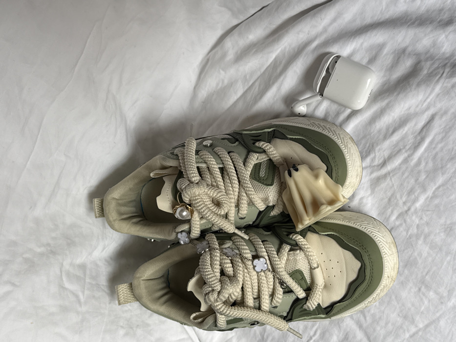 The scene, located in Krasnodar, Russia, depicts a pair of low-top sneakers and an open white Apple AirPods case with earbuds, resting on a white, wrinkled fabric surface. The sneakers are olive green and cream-colored, featuring thick cream-colored laces. The left sneaker's laces are adorned with a silver-colored chain incorporating three white four-leaf clover charms. The right sneaker's laces feature a small gold-colored ring with a pearl-like bead, and a soft, pale yellow ghost-shaped charm is positioned on its toe box. Both sneakers exhibit minor scuffs on their cream-colored soles and include small, clear or silver-colored stud embellishments on their uppers. The AirPods case is situated to the right of the sneakers, open to reveal the two earbuds.