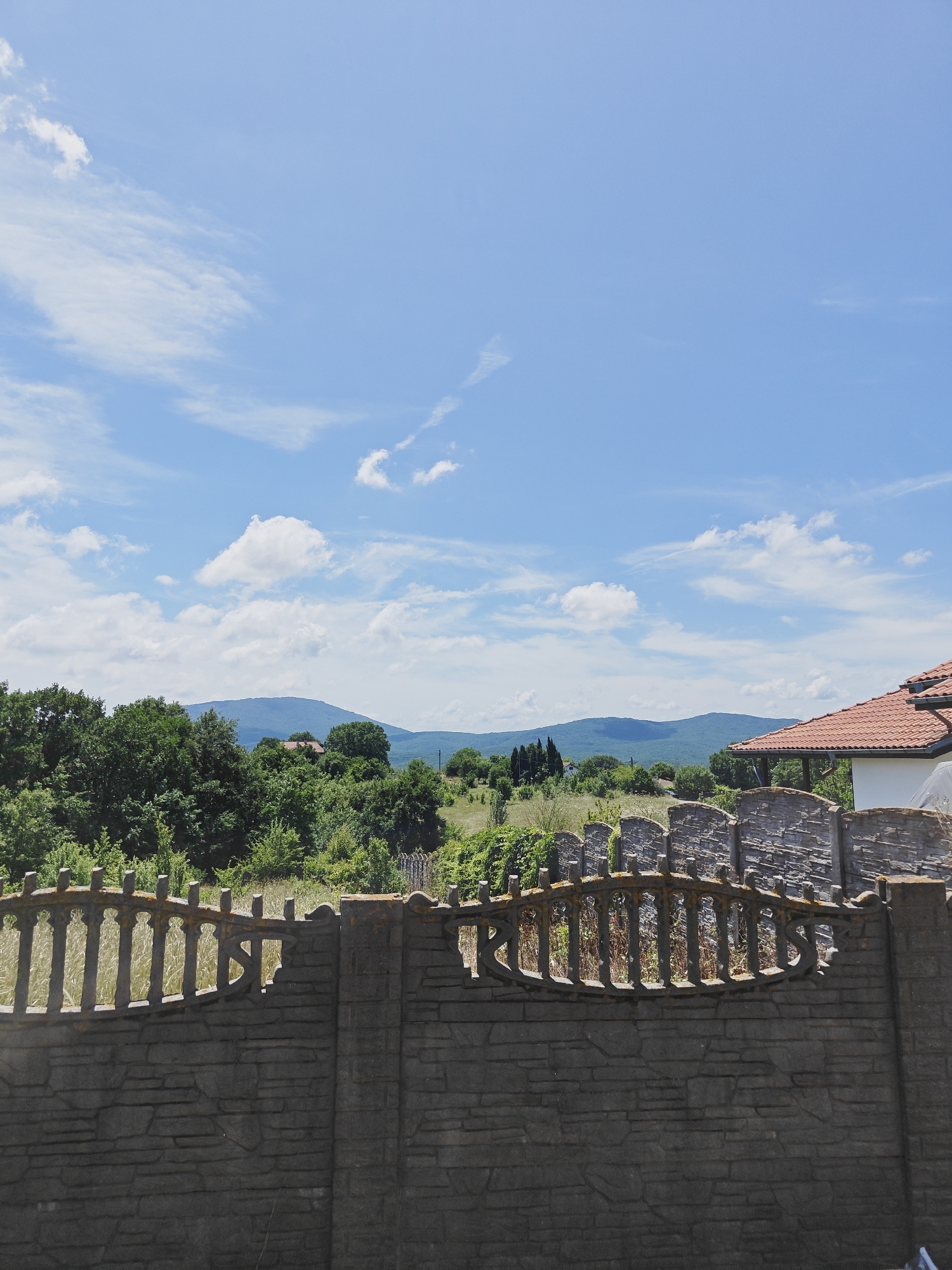 The image depicts a rural landscape in Izgrev, Bulgaria, under a bright blue sky with scattered white clouds. In the foreground, a weathered stone fence with decorative metalwork runs horizontally across the frame. Beyond the fence, a lush green forest covers rolling hills, with a glimpse of a red-tiled roof of a building partially obscured by the trees. In the distance, a range of blue-hued mountains forms the horizon. The scene appears to be captured on a sunny day, suggesting pleasant weather conditions. There are no people or animals visible in the image.