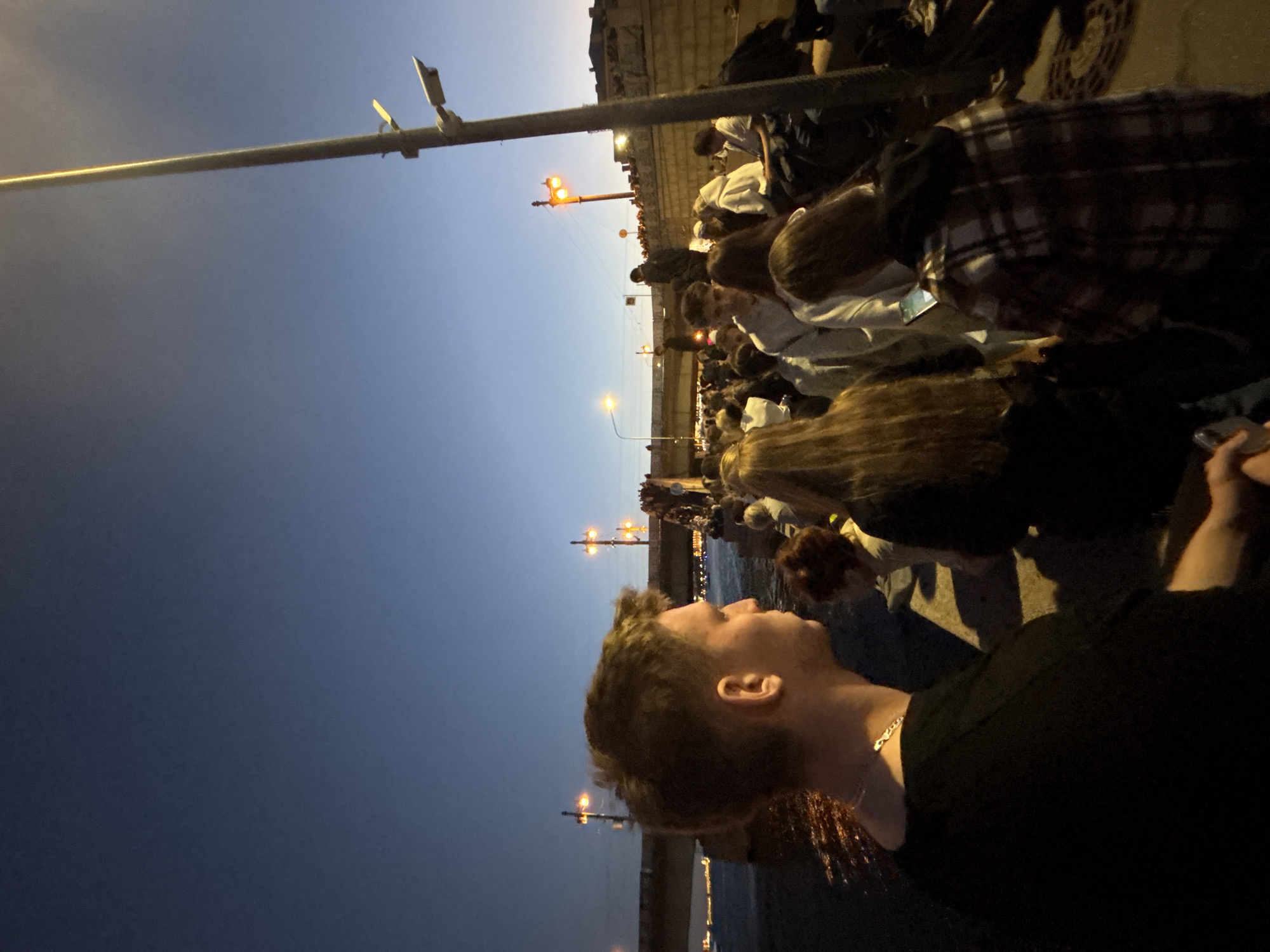 A crowd of people stands near a body of water, possibly the Neva River, in Saint Petersburg, Russia, during twilight. The sky is a deep blue, transitioning from lighter hues near the horizon. A bridge with illuminated arches spans the water in the background, with lights reflecting on the dark surface.
In the foreground, a young man with light brown hair is seen in profile, facing left. He is wearing a black t-shirt and a silver chain necklace. To his right, other individuals are visible, including a woman with long blonde hair and another person wearing a plaid shirt.
A tall lamppost with attached security cameras stands prominently on the right side of the image, casting a faint glow. Streetlights and illuminated structures are scattered throughout the scene, suggesting an urban environment. The overall atmosphere appears to be one of a public gathering or event.
