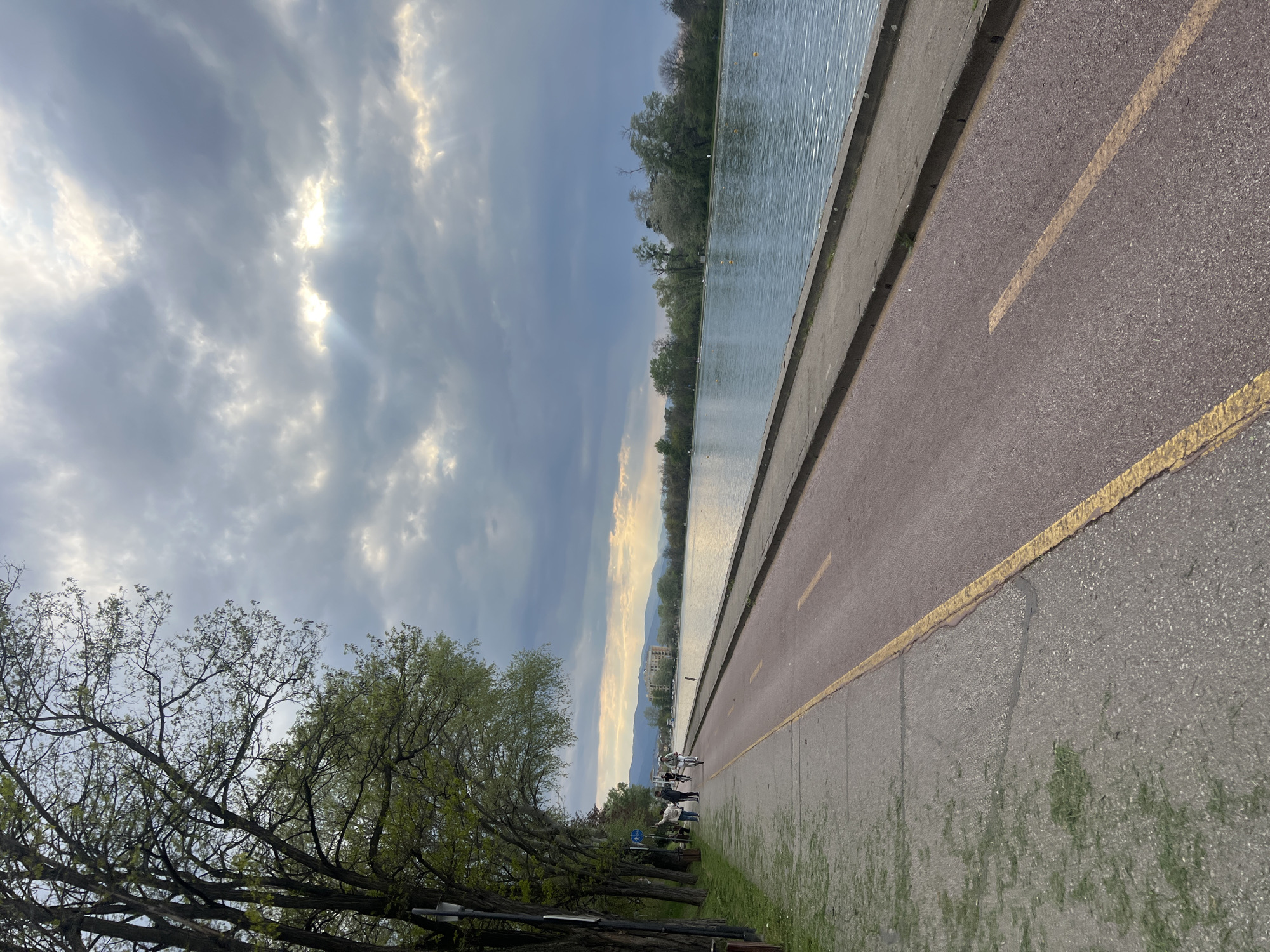 The image captures a serene waterside promenade, likely in Plovdiv, Bulgaria, under a dramatic, partly cloudy sky. The scene depicts a long, calm body of water, possibly a river or canal, stretching into the distance on the right side, its surface reflecting the subdued light. Beyond the water, trees line the far bank, and in the distance, mountains are silhouetted against a sky where soft, golden sunlight pierces through grey clouds, indicating late afternoon or early evening.
On the left, a paved pathway runs parallel to the water. It features a reddish-brown asphalt section, presumably for cyclists, and an adjacent grey concrete section, likely for pedestrians, clearly delineated by a continuous yellow painted line. Alongside the path, a strip of green grass borders a row of mature trees, their branches adorned with fresh, budding green leaves, suggesting a spring setting.
Several people are visible in the middle distance along the pathway, engaging in recreational activities such as walking or cycling, enjoying the pleasant weather. Benches are scattered under the trees, inviting rest. A blue circular sign with a white bicycle symbol is visible on a post, indicating a designated cycling route. In the far distance, across the water, a white multi-story building stands amidst the distant landscape. The overall atmosphere conveys tranquility and leisurely outdoor activity.