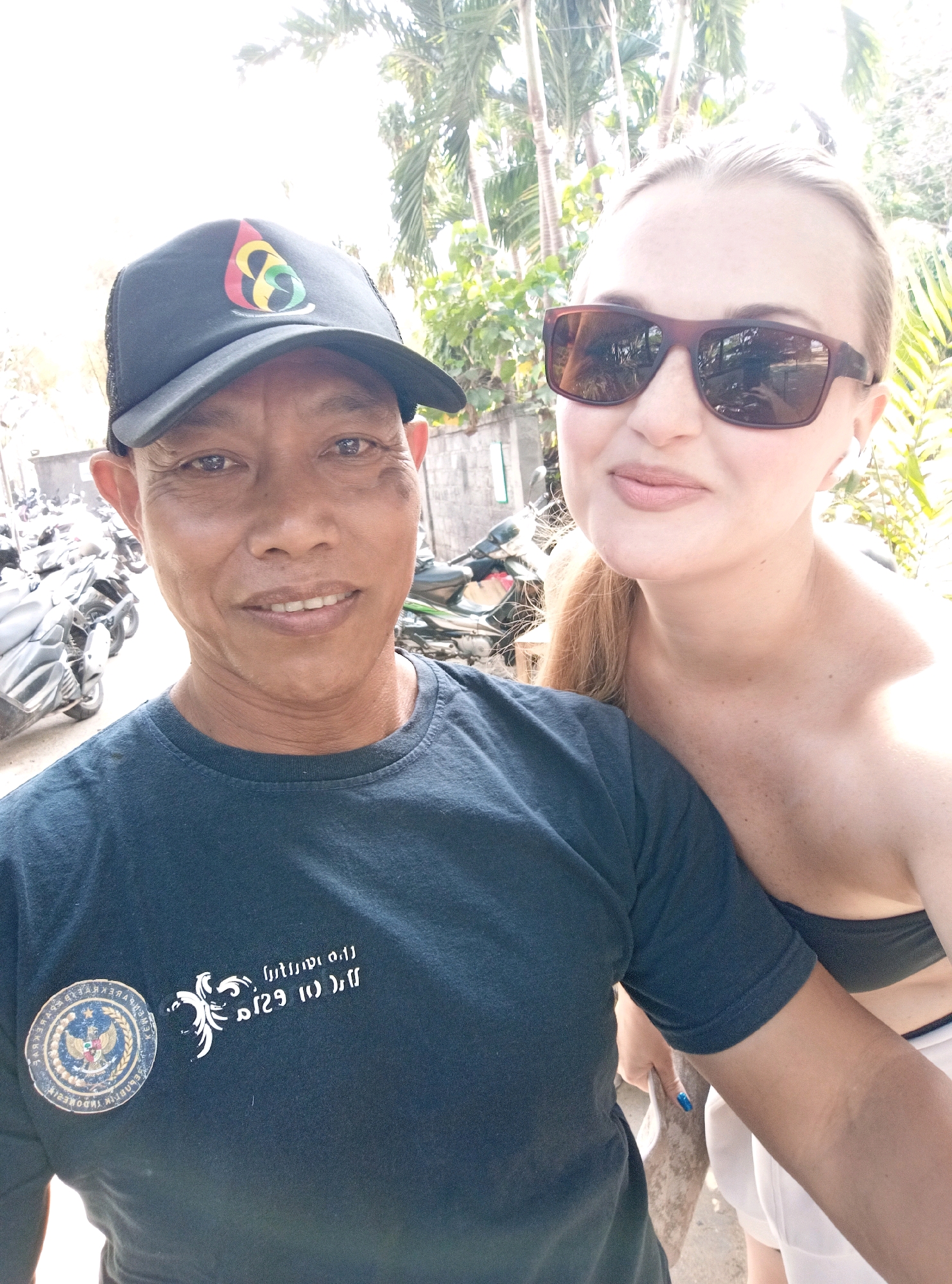A man wearing a dark blue t-shirt and a black baseball cap with a colorful logo stands in the foreground of the image, looking towards the camera. His cap features a logo with a yellow, red, and green design. To his right, a woman with blonde hair and sunglasses is partially visible, also looking towards the camera. She is wearing a strapless top and white bottoms.
The background suggests an outdoor urban environment, possibly in Denpasar, Indonesia. Several motorcycles are parked behind the man, indicating a street or parking area. Palm trees are visible in the background, consistent with a tropical climate. The lighting suggests it is daytime, with bright sunlight casting shadows. The man's t-shirt has a circular emblem on the left chest with what appears to be text around it and an eagle-like crest in the center. There is also a white graphic design on the right side of his shirt. The woman is wearing white wireless earbuds.