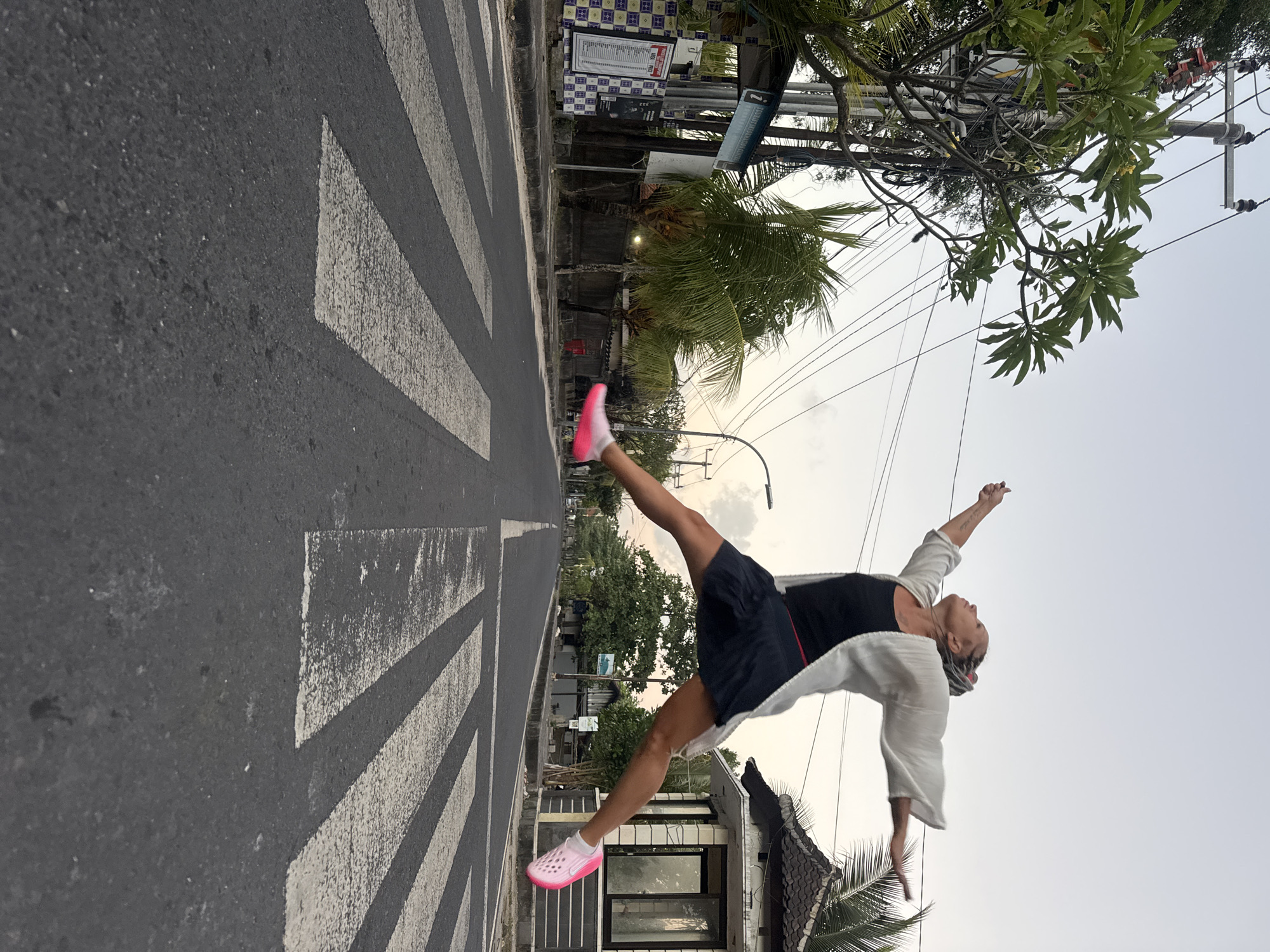 A woman in mid-air, suspended horizontally over a pedestrian crossing, is captured in a full jump. She wears a dark tank top, a short dark skirt, a flowing white open-fronted garment, and bright pink athletic shoes. Her arms are outstretched, her legs are spread, and her body is angled upwards, suggesting a dynamic moment of free flight. The scene unfolds on a street in Kuta, Indonesia, characterized by a road with a clear zebra crossing marking the foreground. Utility poles with an entanglement of wires stretch across the sky, and lush green trees, including palm trees, line the sides of the road, interspersed with buildings and what appears to be a small kiosk or booth on the right. The lighting suggests dusk or early morning, with a soft, diffused sky.