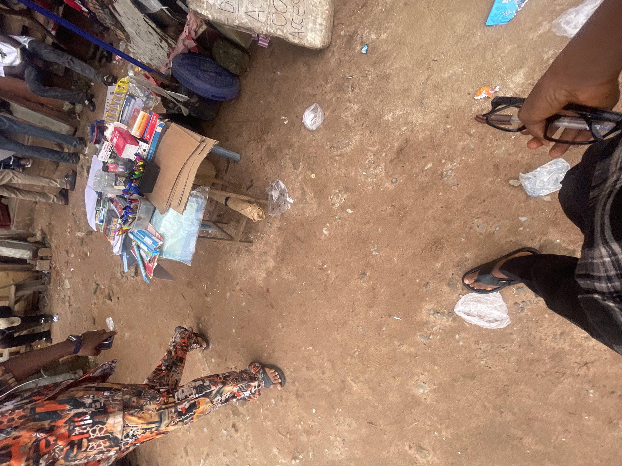 The image depicts an outdoor scene in Ihiagwa, Nigeria, likely a market or roadside stall. In the foreground, a person wearing black pants and a plaid shirt is seen from the waist down, holding a pair of glasses. To their left, a person in vibrantly patterned pants and sandals is standing. In the mid-ground, a table is set up with various stationery items, including pens, notebooks, and boxes. Several people are visible in the background, some standing and others sitting. The ground is composed of reddish-brown dirt with scattered debris, including plastic bags and paper. A blue plastic container and other objects are visible near the stall. The overall impression is one of a bustling, informal commercial environment.