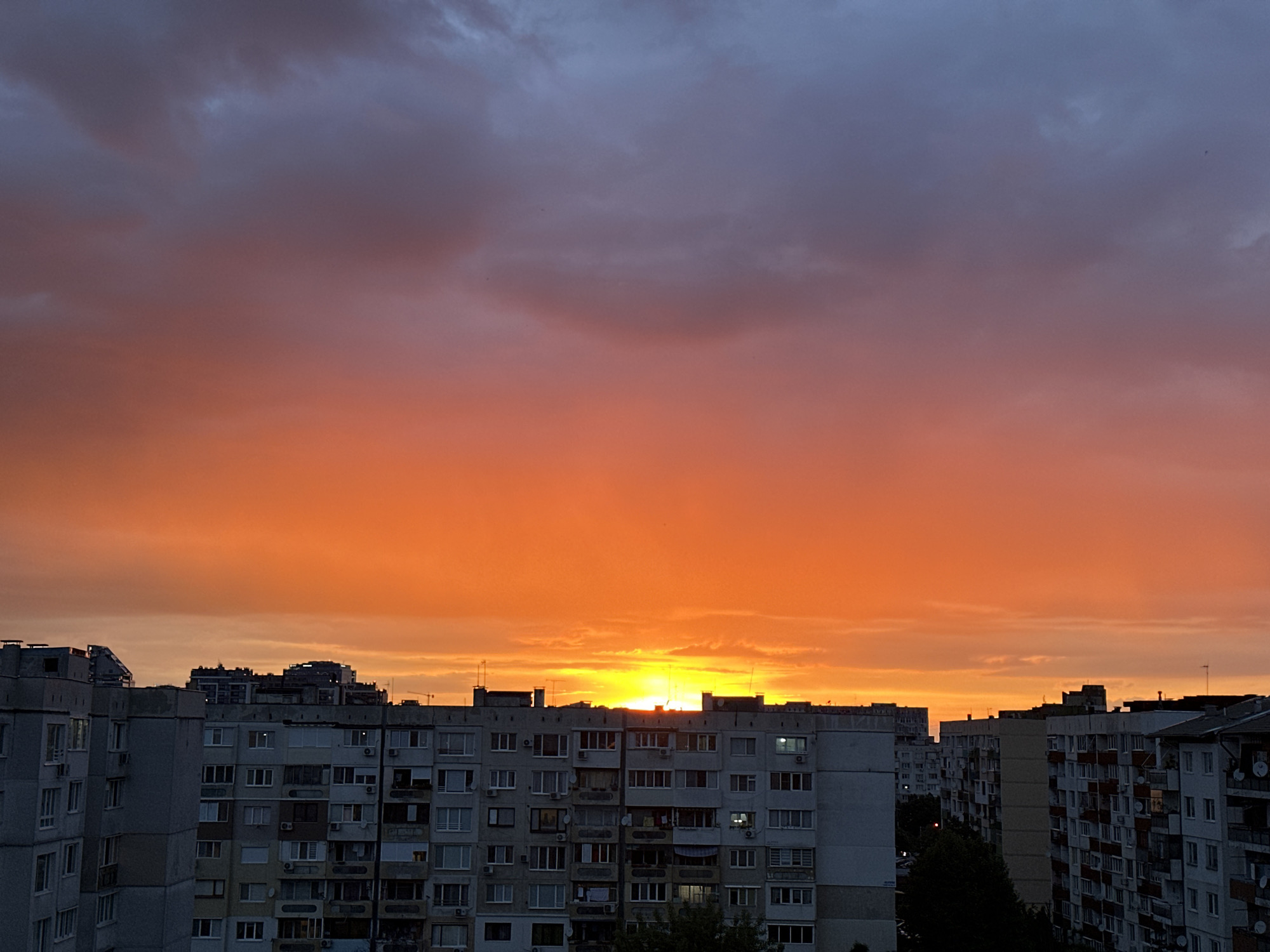 The image captures a vibrant sunset over the urban landscape of Sofia, Bulgaria. A wide expanse of orange and red hues dominates the sky, transitioning into darker shades of gray and blue at the top. The sun is setting below the horizon, its light peeking through the gaps between the buildings. Below the sky, a cluster of residential apartment buildings fills the lower portion of the image. These are typical Soviet-era concrete block structures, characterized by their rectangular shapes and rows of windows. A few trees are visible between the buildings. There are no people visible in the scene. The scene conveys a sense of urban life amidst the beauty of a sunset.