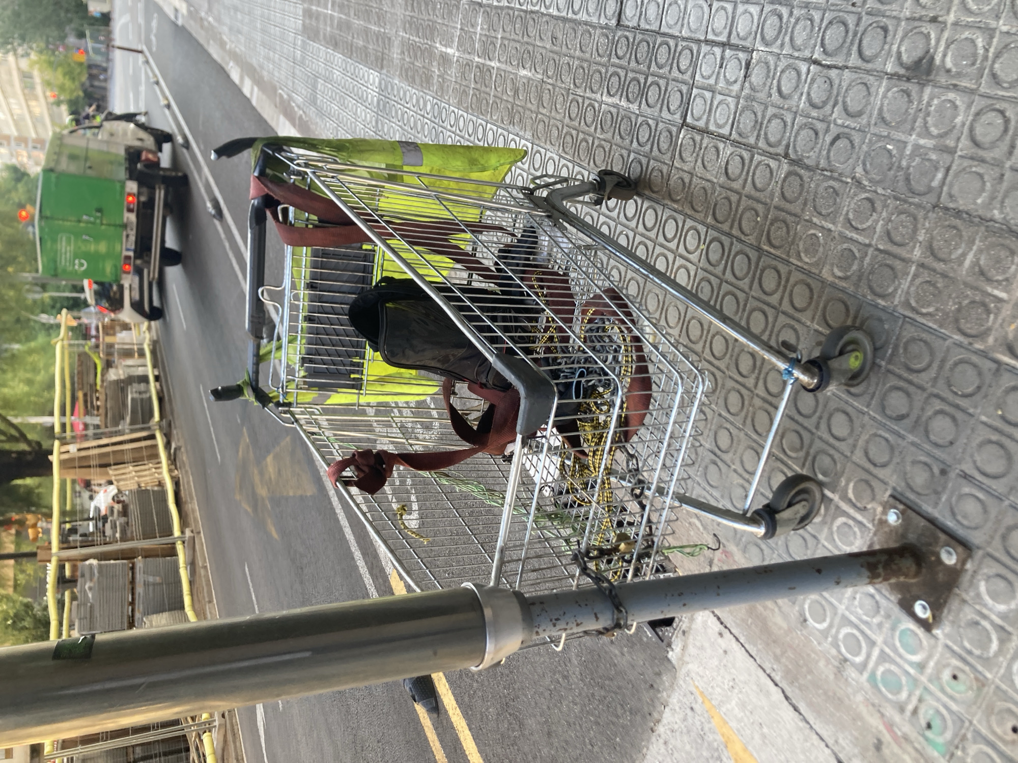 This media file captures a scene on a street in Barcelona, Spain. The primary subject is a shopping cart, positioned on a textured sidewalk with a repeating circular pattern. The shopping cart is filled with various items, including a black bag, some red straps, and a bright green piece of fabric. Yellow strings are also visible within the cart.
In the background, a street is visible, with a green truck parked on the left side. The truck appears to be a utility or service vehicle. Further down the street, there is construction or renovation work in progress, with stacks of materials and scaffolding. The scene suggests a daytime setting, likely in fair weather, given the clarity of the image and the general appearance of the lighting. The presence of a shopping cart on the sidewalk and the construction activity indicate an urban environment. There are no visible people clearly in focus, but the truck and the construction site suggest some human activity is present or has recently occurred. The overall mood is observational, documenting a slice of urban life.