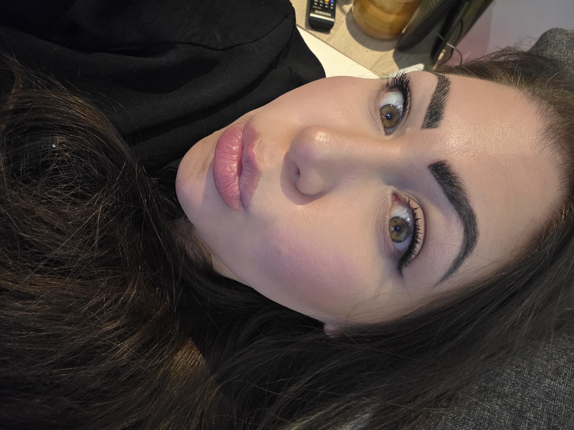 A close-up, angled portrait depicts a young woman with fair skin, prominent cheekbones, and full lips wearing pink lipstick. She has thick, dark eyebrows and hazel eyes with dark eyeliner and mascara. Her dark brown hair is visible around her face and shoulder. She appears to be lying down. In the upper portion of the frame, background objects include a remote control and what seems to be a jar. The image does not provide any indications of location in London, United Kingdom, and there are no actions, interactions, or environmental details that can be extracted.