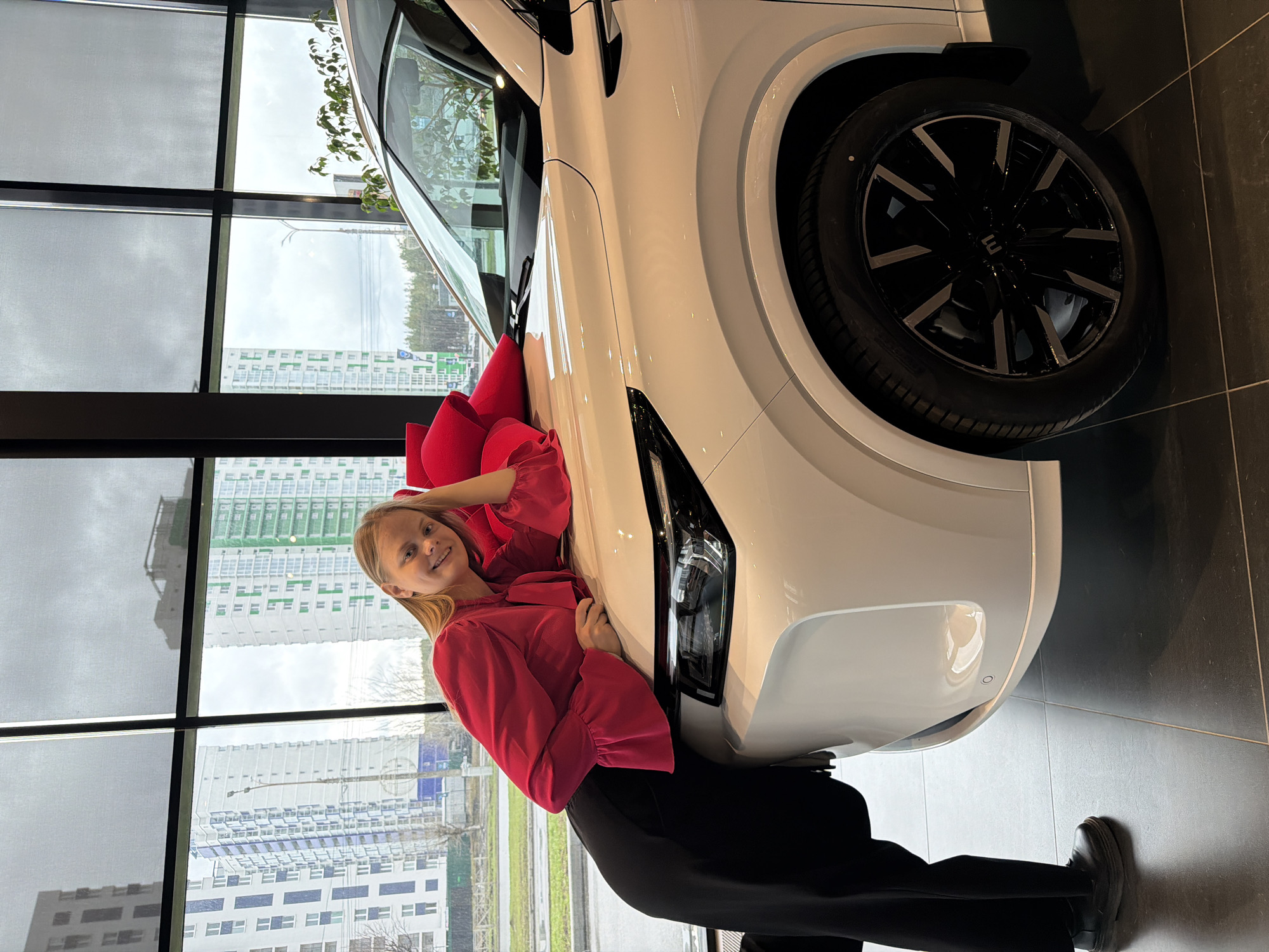 A young woman in a red blouse and black pants poses with a new white car, adorned with a large red bow, inside a car dealership. The background features a large window showing an urban cityscape of Izhevsk, Russia. Multiple multistory buildings are visible outside the window. The car is parked on a dark tile floor.