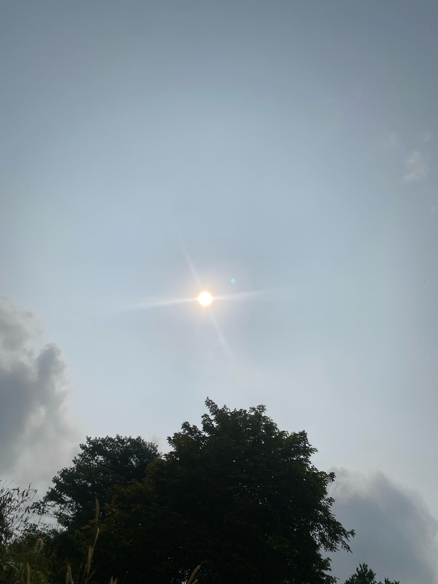 The image shows a sky with the sun shining brightly, creating a lens flare effect. Dark clouds are present on the left side of the frame, and lighter clouds are scattered throughout. The tops of several trees are visible at the bottom of the image, silhouetted against the sky. The scene appears to be outdoors, likely in a natural setting, with no specific urban elements visible. The location is described as Unknown, Unknown.