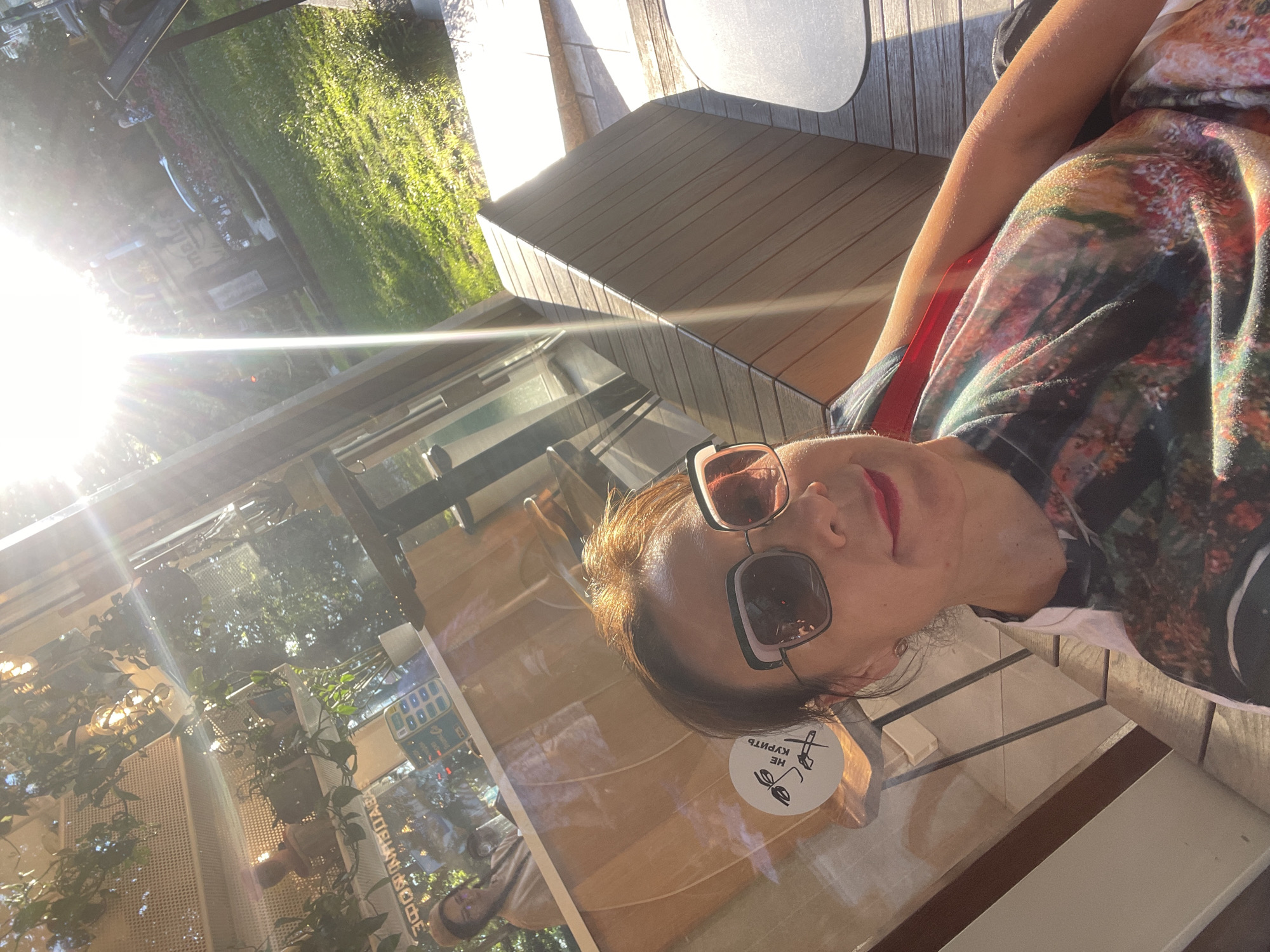 In the city of Sochi, Russia, a woman is captured in a selfie, her face framed by stylish sunglasses. She is seated outdoors, likely at a cafe or restaurant, judging by the presence of tables and chairs visible in the background. The bright sunlight, a common feature of Sochi's climate, creates a lens flare effect across the image, suggesting it was taken on a clear, sunny day. The woman's attire consists of a colorful, patterned t-shirt, and she is wearing lipstick. In the background, through a glass window, another person is visible, seemingly working behind a counter. The environment suggests a relaxed, possibly urban setting with greenery and outdoor seating.