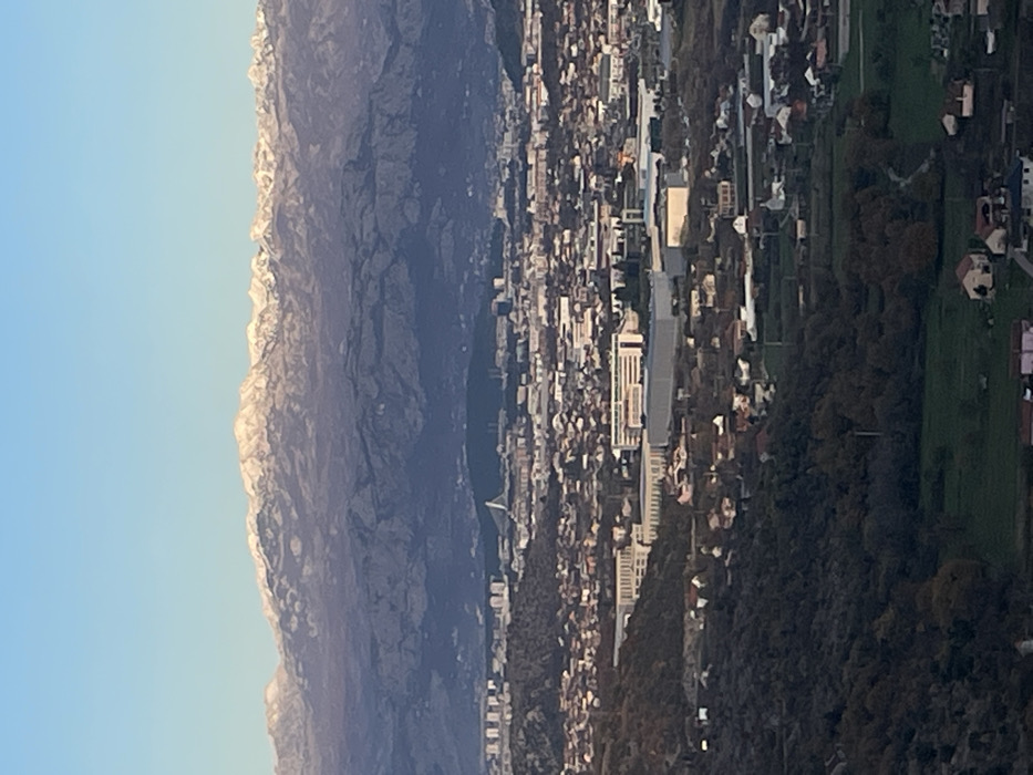 A panoramic, high-angle view depicts an urbanized valley landscape under a clear blue sky, extending towards snow-capped mountains in the distant background. The scene is located in Montenegro, overlooking the area encompassing Gornji Kokoti and the city of Podgorica.
The upper fifth of the image is occupied by a clear, light blue sky. Below this, a range of rugged mountains, dark in tone, exhibits patches of white snow on its peaks and upper slopes.
The mid-ground features a sprawling urban and suburban environment. In the further mid-distance, a denser city, identifiable as Podgorica, is visible with numerous high-rise buildings and a prominent white cable-stayed bridge (Millennium Bridge) spanning a river. Closer to the foreground, the terrain consists of rolling hills and valley floor, developed with a mix of residential structures, characterized by red and light-colored roofs, interspersed with green plots of land and scattered trees. This area represents the locale of Gornji Kokoti, extending into the suburban sprawl leading to Podgorica. Larger, rectangular commercial or industrial buildings with flat roofs are also visible within this mid-ground urban fabric.
The immediate foreground, occupying the bottom fifth of the image, shows dark, heavily vegetated hillside terrain, indicating the elevated vantage point from which the photograph was taken. A few isolated houses with visible roofs are situated on these lower slopes.
No individuals, specific actions, or interactions are discernible due to the distant, high-altitude perspective. The lighting suggests either late afternoon or early morning, casting long shadows across parts of the landscape and highlighting the snow on the distant mountains.