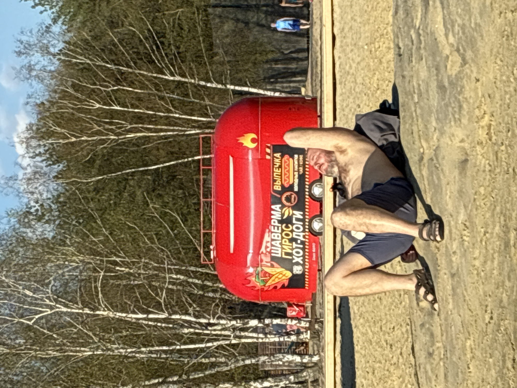 The image shows a man sitting on the ground next to a red food truck with Russian writing on it. The truck advertises "SHAWERMA" and "HOT-DOGI" along with images of food items and flames. The man is shirtless, wearing dark shorts and sandals. He has his hand on his head. The location appears to be outdoors, possibly a park or recreational area, with birch trees in the background. In the distance, there is another person walking. The setting is in an unknown location in the city of Unknown, Unknown.
