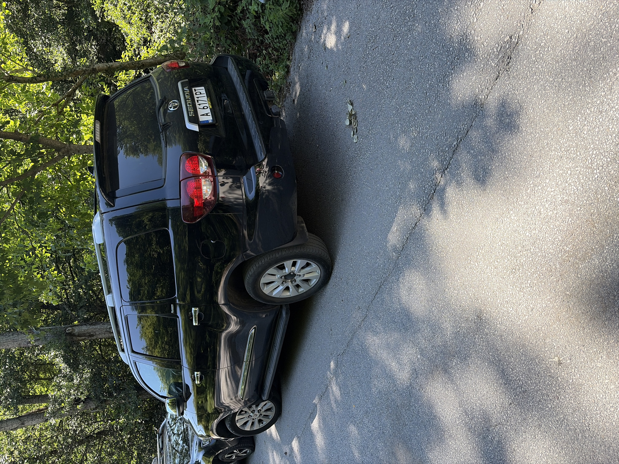 A black Toyota Sequoia SUV is parked on an asphalt road, partially shaded by trees. The SUV's license plate reads "A 6171 PT" and "IMPORTING." To the left, another dark SUV is partially visible. The environment is wooded, with sunlight filtering through the leaves, casting dappled shadows on the road. The scene appears to be on a quiet road in the city of Devin, Bulgaria.