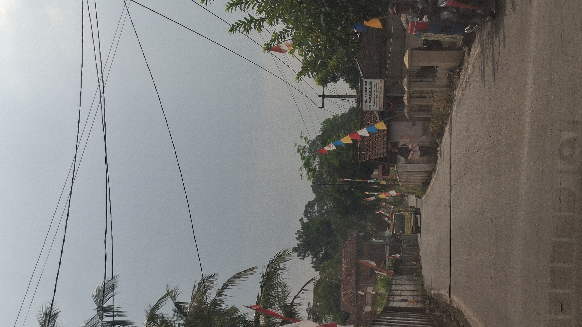 The image shows a street scene in Sirnagalih, Indonesia. The sky is overcast. Power lines are visible overhead. On the left side of the road, there are houses with tiled roofs and a concrete wall. Indonesian flags are displayed along the road. On the right side of the road, there are more houses with tiled roofs, a sign that reads "PRATEX MANDIRI BIDAR", and a tree with some flags. A yellow truck is driving down the road. A person in a headscarf and dark pants is standing on the right side of the road. Further down the road on the right, a person is sitting on a motorcycle. The road is paved and has a white line dividing it.