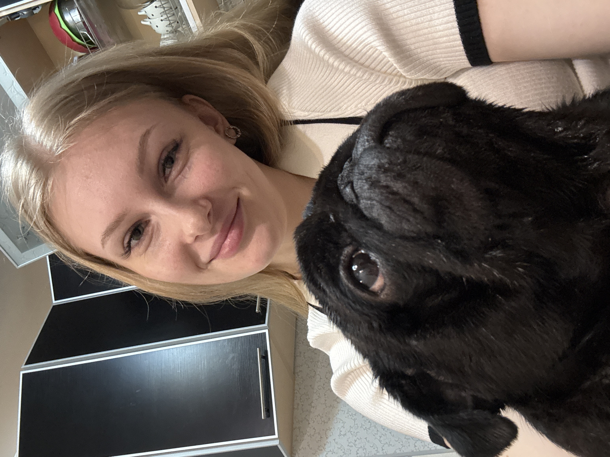 A young, blonde-haired woman with light skin and earrings is taking a selfie with a black pug. The woman is wearing a white, ribbed sweater with black trim on the sleeves. The dog is in the foreground, close to the camera, its face filling the right side of the frame. The woman is smiling slightly, and the dog is looking at the camera. The background includes black and silver kitchen cabinets and a glimpse of a dish rack. The image was likely taken indoors in a residential kitchen. The city of Perm, Russia, is not identifiable from the image alone.