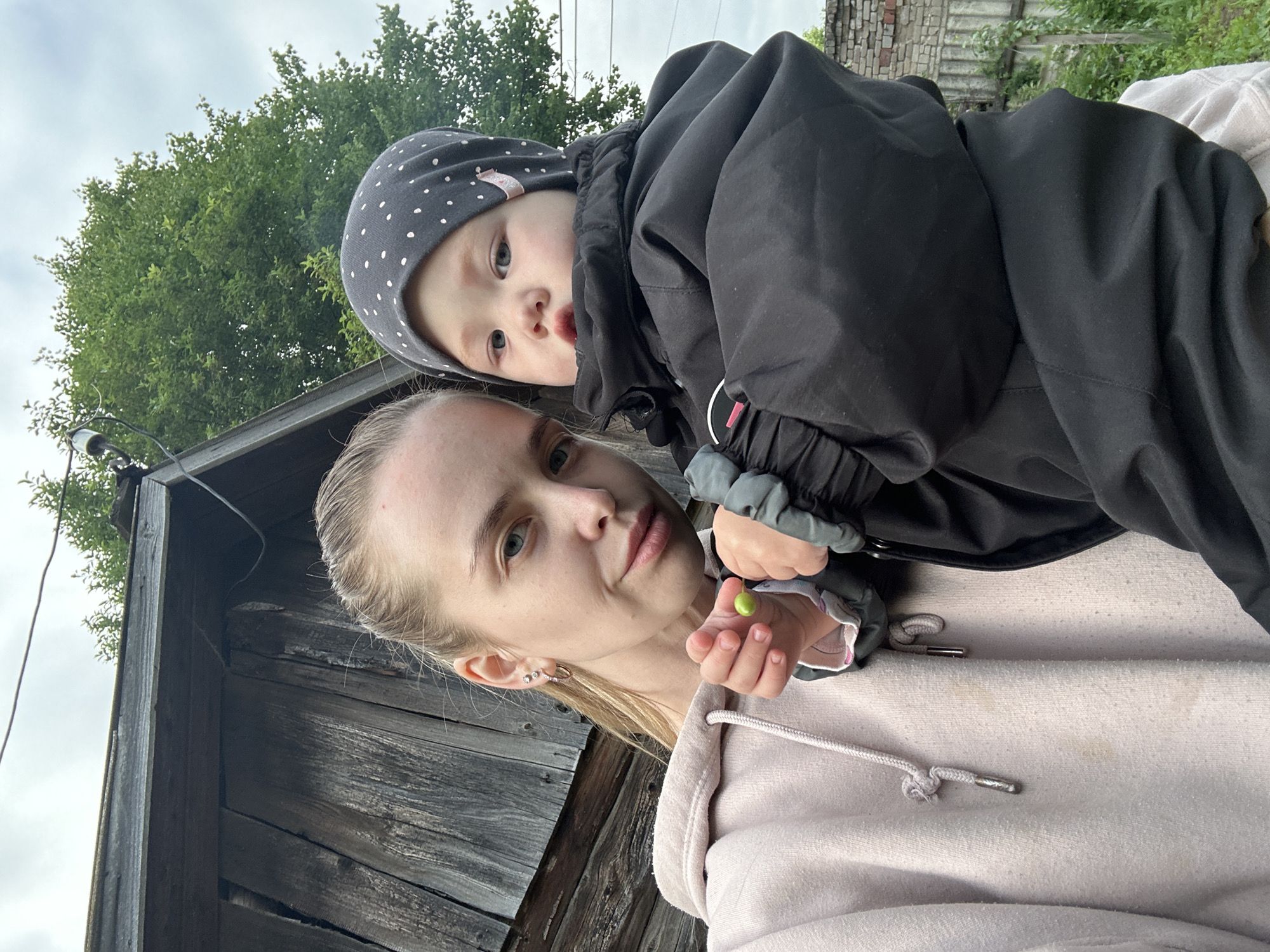 In the city of Azinsky, Russia, a young Caucasian woman with blonde hair tied back in a ponytail is holding a baby. The woman is wearing a light pink hooded sweatshirt. The baby is wearing a grey dotted beanie and a black jacket. The background consists of an old wooden structure, green trees, and an overcast sky. The woman is holding a small green object in her left hand.