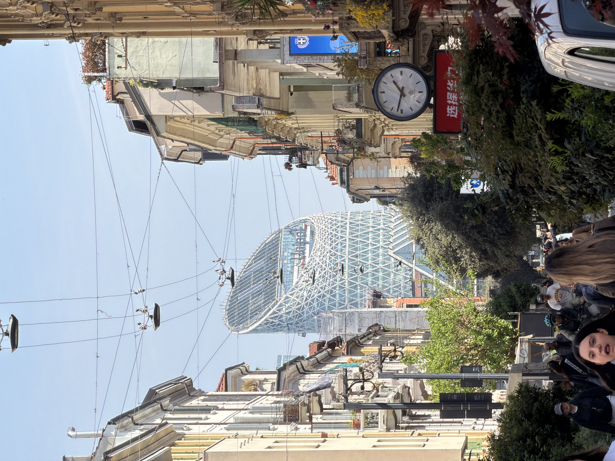 The image shows a street scene in Milan, Italy, looking towards a modern architectural building. The street is lined with older buildings with ornate facades, some showing signs of age. Overhead electrical lines crisscross the street. A clock hangs on one of the buildings, and a red sign with Chinese characters is visible nearby. A group of people is on the right side of the image, suggesting a sidewalk or pedestrian area. The focal point is the contemporary building, which has a distinctive, curved, latticework design. The sky is visible above the buildings.