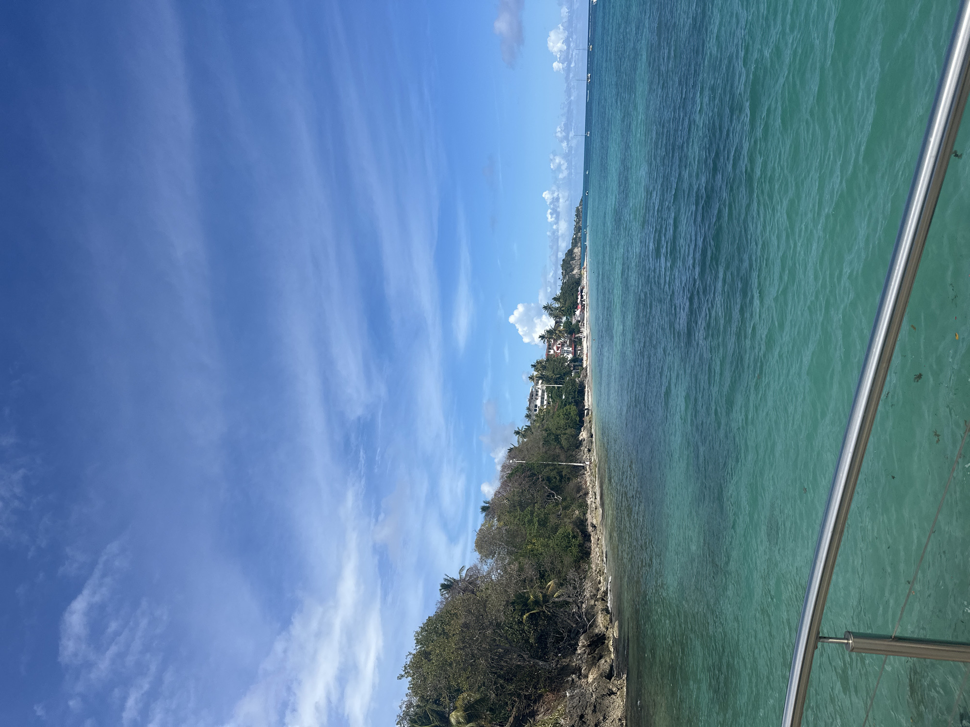 Captured from an elevated perspective, possibly a boat or cliff edge, the image showcases a vibrant coastal scene near the city of Le Gosier, France. The right of the frame features turquoise waters, with a metallic railing from the vantage point cutting into the scene. Along the shoreline, lush green vegetation extends to the edge of rocky terrain. Further inland, buildings are visible amidst the trees, indicating a settled area. Above, a striking blue sky filled with linear, wispy clouds dominates the left half of the image.