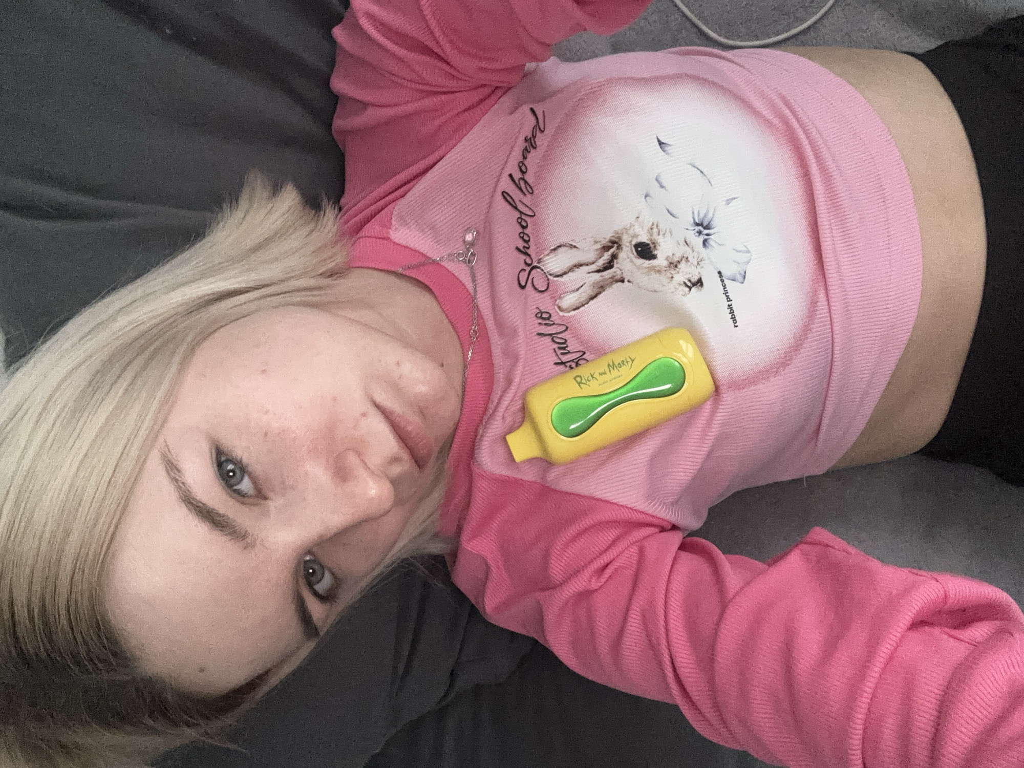 A young woman with fair skin and blond hair is lying down on a gray surface. She is wearing a pink long-sleeved shirt with a graphic design featuring a rabbit and the words "Radio School Board" and "rabbit princess." A yellow and green object, appearing to be a bottle with "Rick and Morty" printed on it, is positioned on her chest. She has blue eyes and is looking directly at the camera. The location is unknown, and there is no indication of Russia or any city.