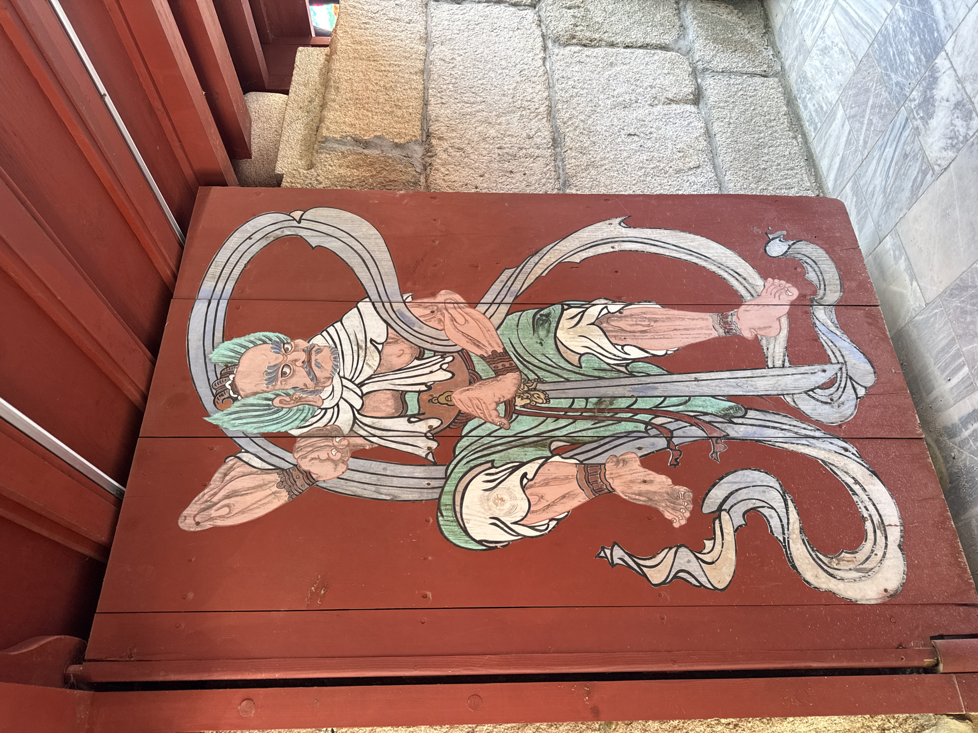 The image depicts a painted wooden panel with a figure of a deity in a stylized pose. The figure has a long white beard, light green hair, and is adorned with flowing white garments and green robes. It appears to be floating or reclining, with its limbs positioned in a dynamic fashion, holding a sword in its hands. The background of the panel is red, made up of horizontal wooden planks.
The panel is located in a Korean Buddhist temple in Seoul, South Korea, specifically at Jogyesa Temple (조계사). It serves as a depiction of one of the guardians of Buddhism. The panel is mounted on a wooden frame, adjacent to a stone-paved area and a portion of the red-painted wooden architecture of the temple.