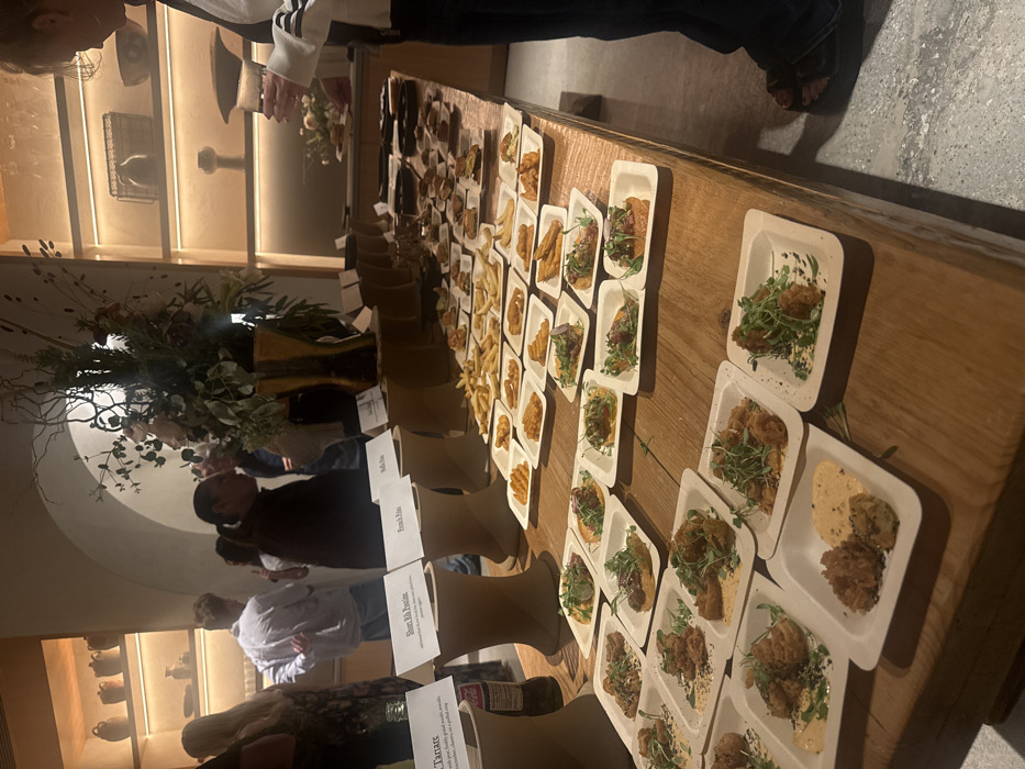 The image displays an indoor food event or tasting taking place in Los Angeles, United States of America. A long, rustic wooden table, extending from the lower left to the upper right of the frame, is covered with numerous individual portions of appetizers. Each portion is presented on a small, square, off-white plate, likely disposable.
Identifiable food items, arranged in neat rows, include piles of French fries, multiple plates of fried items with sauces and green garnish (possibly fried chicken or seafood), and bruschetta-style toppings on bread. White tent cards placed on elevated stands identify some dishes, with visible labels for "Tartare", "Short Rib Poutine", and "French Fries". A large floral arrangement featuring branches and light-colored flowers in a gold-toned vase stands on the table towards the center-back. A partial Coca-Cola bottle is visible on the far left.
In the background, several individuals are present, appearing blurred due to shallow depth of field, indicating a social gathering. On the far right, a person wearing dark pants and a white long-sleeved top with black stripes on the sleeves is partially visible, holding a clear drinking glass in their left hand and reaching towards the food. Another person with dark hair pulled into a ponytail is seen in the mid-ground, observing the scene. The ambient lighting is warm and indirect, illuminating decorative shelves on the back wall which display various pottery and objects. The floor appears to be a light-colored concrete surface.